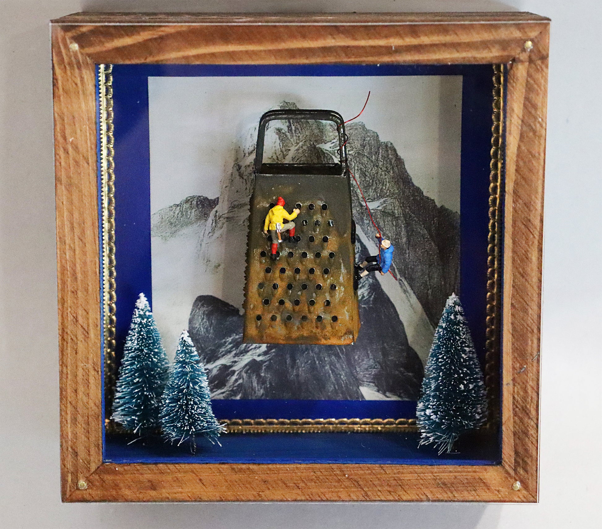 Original Objekt - Assemblage von Elisabeth Dishur, Titel „Der Bergsteiger“ – Objektkiste mit Miniatur-Figuren: Zwei Bergsteiger erklimmen eine Küchenreibe, darunter 3 verschneite Tannen, im Hintergrund eine Bergspitze. Eingefasst in eine Holzkiste, mit Plexiglas versehen - Objektcollage, erhältlich bei Galerie Waldbach - What The Artist Made