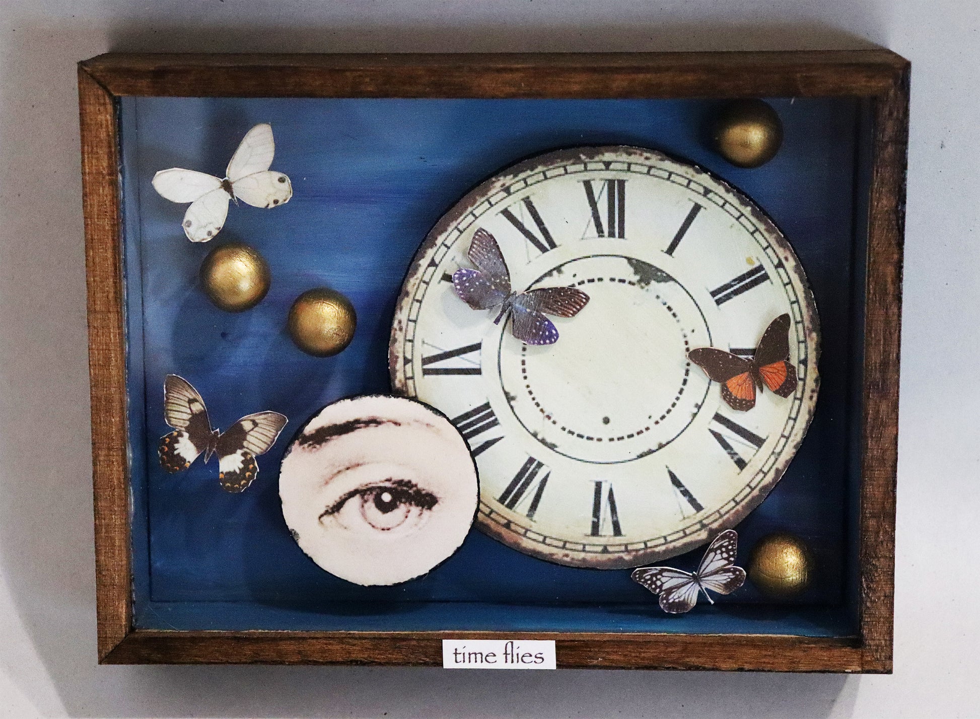 Original Objekt - Assemblage von Elisabeth Dishur, Titel „time flies“ – Objektkiste mit Papiercollagen und Gegenständen: Ein Ziffernblatt aus Papier, das die Kiste dominiert, daneben aus Papier ein rundes  Auge. Fünf Schmetterlinge aus Papier und 4 goldene Kugeln sind darum angeordnet, der Hintergrund ist blau und der Titel steht unten am Rand auf dem Holz. Eingefasst in eine Holzkiste, mit Plexiglas versehen - Objektcollage, erhältlich bei Galerie Waldbach - What The Artist Made