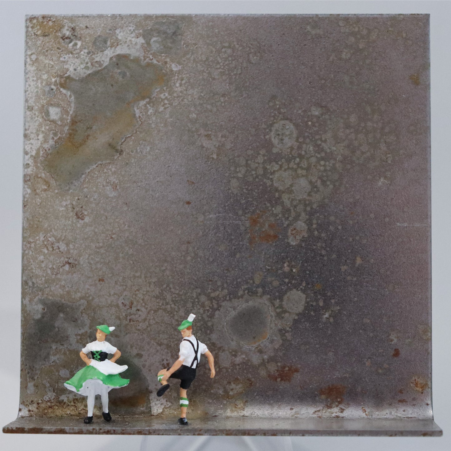 Original Objekt - Assemblage von Katrin Schramm, Titel „Auf der Balz...“ – Metallwinkel mit Miniatur-Figuren: Ein mann und eine Frau in grün-weißen bayrischen Trachten tanzen auf der linken Seite des Rahmens. Dahinter das Metall des Winkels mit Struktur, als Hintergrund. Objektcollage, erhältlich bei Galerie Waldbach - What The Artist Made
