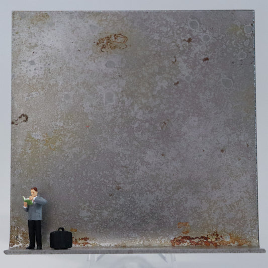 Original Objekt - Assemblage von Katrin Schramm, Titel „Bücher sind Lebensmittel“ – Metallwinkel mit Miniatur-Figur: Ein Mann steht am linken Rand des Winkels und liest in einem Buch. Neben ihm steht seine Tasche. Dahinter das Metall des Winkels mit Struktur, als Hintergrund. Objektcollage, erhältlich bei Galerie Waldbach - What The Artist Made