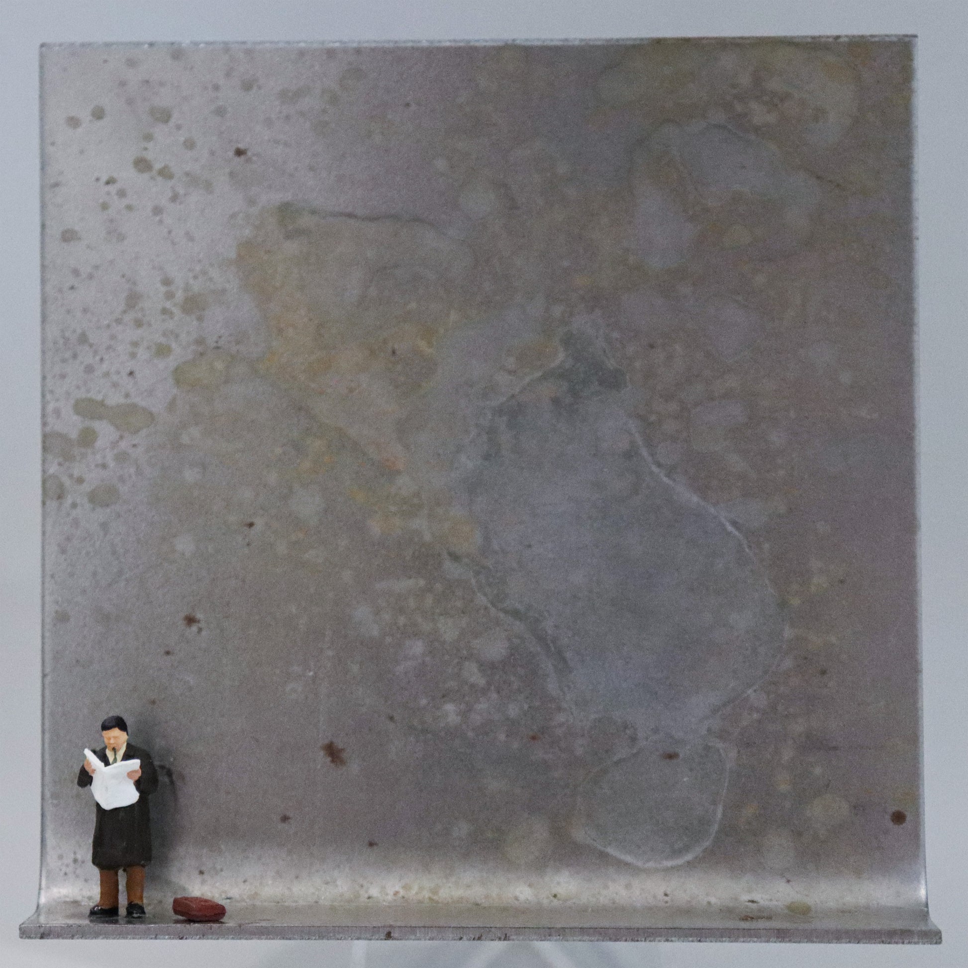 Original Objekt - Assemblage von Katrin Schramm, Titel „Der letzte Zeitungsleser?“ – Metallwinkel mit Miniatur-Figur: Ein Mann steht am linken Rand des Winkels und liest in einer aufgeschlagenen Zeitung. Neben ihm liegt  seine Tasche. Dahinter das Metall des Winkels mit Struktur, als Hintergrund. Objektcollage, erhältlich bei Galerie Waldbach - What The Artist Made