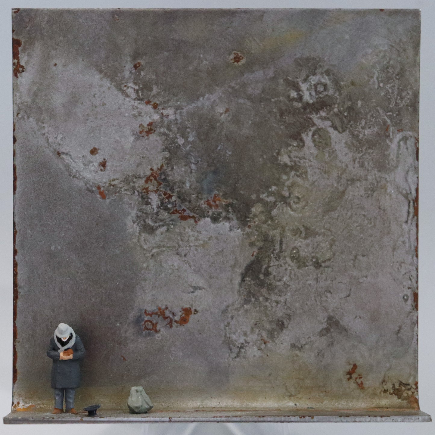 Original Objekt - Assemblage von Katrin Schramm, Titel „Geben...“ – Metallwinkel mit Miniatur-Figur: Ein Mann steht am linken Rand des Winkels in gebeugter Haltung. Er hält die Hände zur Brust, neben ihm liegt ein Hut auf dem Boden mit der Öffnung nach oben. Daneben steht ein großer Rucksack. Dahinter das Metall des Winkels mit Struktur, als Hintergrund. Objektcollage, erhältlich bei Galerie Waldbach - What The Artist Made
