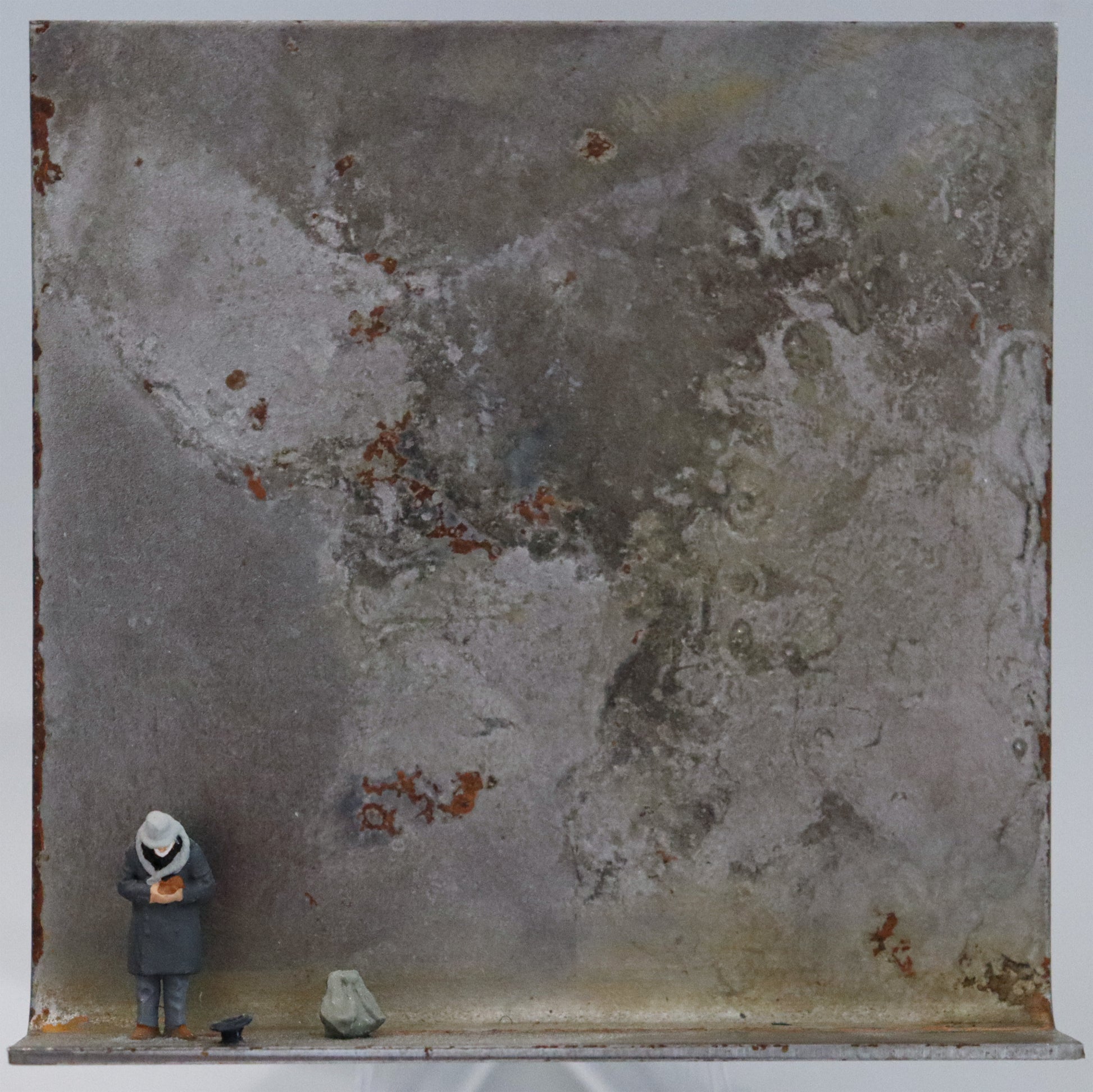 Original Objekt - Assemblage von Katrin Schramm, Titel „Geben...“ – Metallwinkel mit Miniatur-Figur: Ein Mann steht am linken Rand des Winkels in gebeugter Haltung. Er hält die Hände zur Brust, neben ihm liegt ein Hut auf dem Boden mit der Öffnung nach oben. Daneben steht ein großer Rucksack. Dahinter das Metall des Winkels mit Struktur, als Hintergrund. Objektcollage, erhältlich bei Galerie Waldbach - What The Artist Made