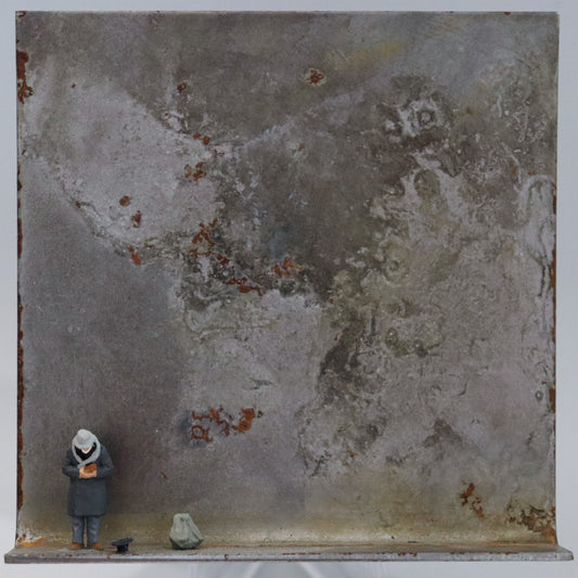 Original Objekt - Assemblage von Katrin Schramm, Titel „Geben...“ – Metallwinkel mit Miniatur-Figur: Ein Mann steht am linken Rand des Winkels in gebeugter Haltung. Er hält die Hände zur Brust, neben ihm liegt ein Hut auf dem Boden mit der Öffnung nach oben. Daneben steht ein großer Rucksack. Dahinter das Metall des Winkels mit Struktur, als Hintergrund. Objektcollage, erhältlich bei Galerie Waldbach - What The Artist Made