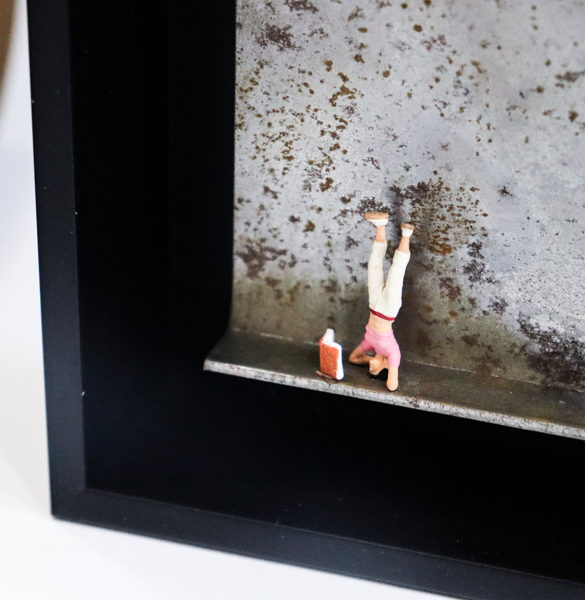 Original Objekt - Assemblage von Katrin Schramm, Titel „180°“ – Metallwinkel mit Miniatur-Figur, Detailansicht:  Eine Frau macht einen Kopfstand, vor ihr steht ein Buch aufgeklappt. Dahinter das Metall des Winkels mit Struktur, als Hintergrund. In einem schwarzen Rahmen aus Holz. Objektcollage, erhältlich bei Galerie Waldbach - What The Artist Made