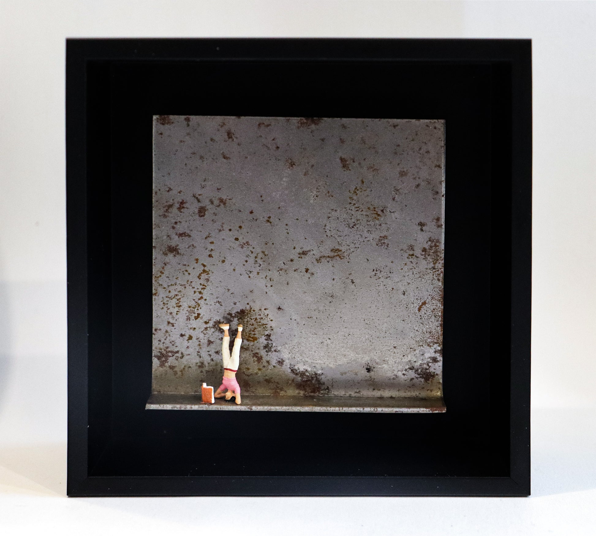 Original Objekt - Assemblage von Katrin Schramm, Titel „180°“ – Metallwinkel mit Miniatur-Figur:  Eine Frau macht einen Kopfstand, vor ihr steht ein Buch aufgeklappt. Dahinter das Metall des Winkels mit Struktur, als Hintergrund. In einem schwarzen Rahmen aus Holz. Objektcollage, erhältlich bei Galerie Waldbach - What The Artist Made