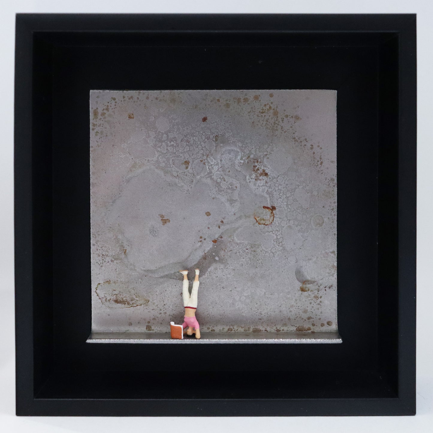 Original Objekt - Assemblage von Katrin Schramm, Titel „180 °“ – Metallwinkel mit Miniatur-Figur: Eine Frau macht einen Kopfstand. Vor ihr steht ein Buch, das sie währenddessen liest. Dahinter das Metall des Winkels mit Struktur, als Hintergrund. Objektcollage im schwarzen Rahmen, erhältlich bei Galerie Waldbach - What The Artist Made
