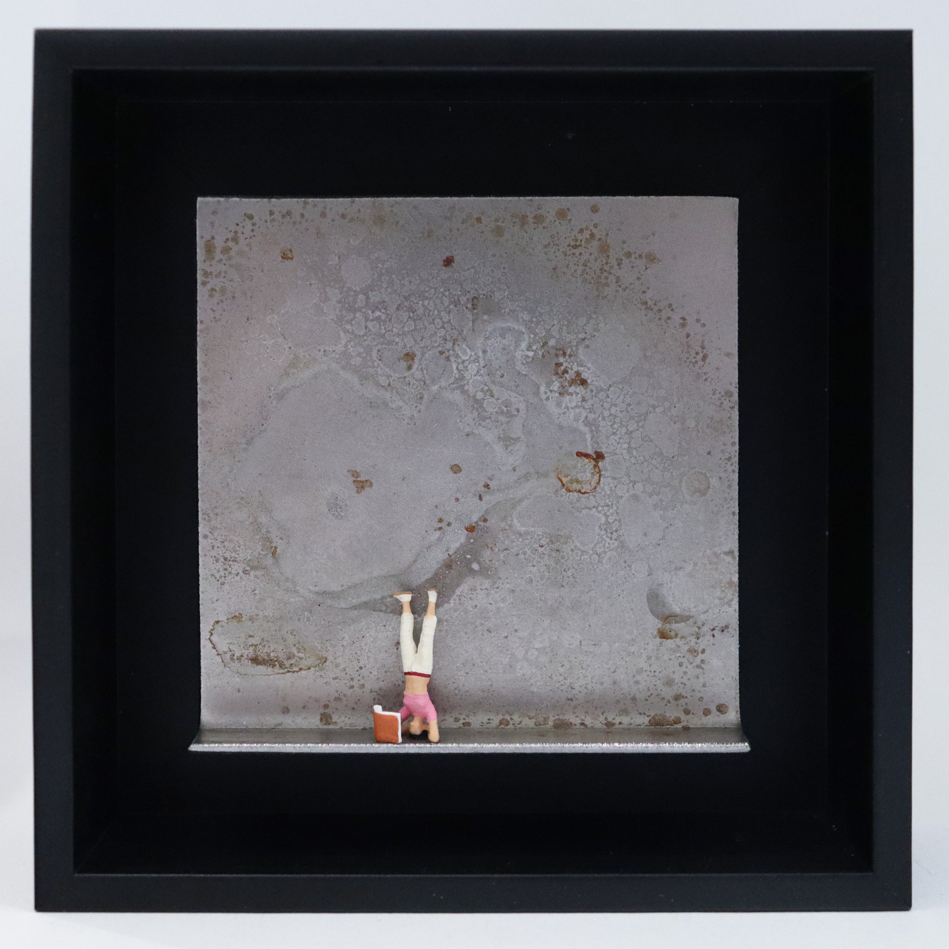 Original Objekt - Assemblage von Katrin Schramm, Titel „180 °“ – Metallwinkel mit Miniatur-Figur: Eine Frau macht einen Kopfstand. Vor ihr steht ein Buch, das sie währenddessen liest. Dahinter das Metall des Winkels mit Struktur, als Hintergrund. Objektcollage im schwarzen Rahmen, erhältlich bei Galerie Waldbach - What The Artist Made