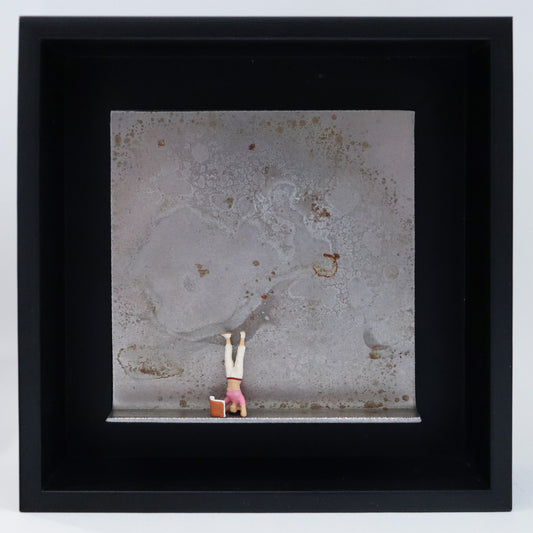 Original Objekt - Assemblage von Katrin Schramm, Titel „180 °“ – Metallwinkel mit Miniatur-Figur: Eine Frau macht einen Kopfstand. Vor ihr steht ein Buch, das sie währenddessen liest. Dahinter das Metall des Winkels mit Struktur, als Hintergrund. Objektcollage im schwarzen Rahmen, erhältlich bei Galerie Waldbach - What The Artist Made