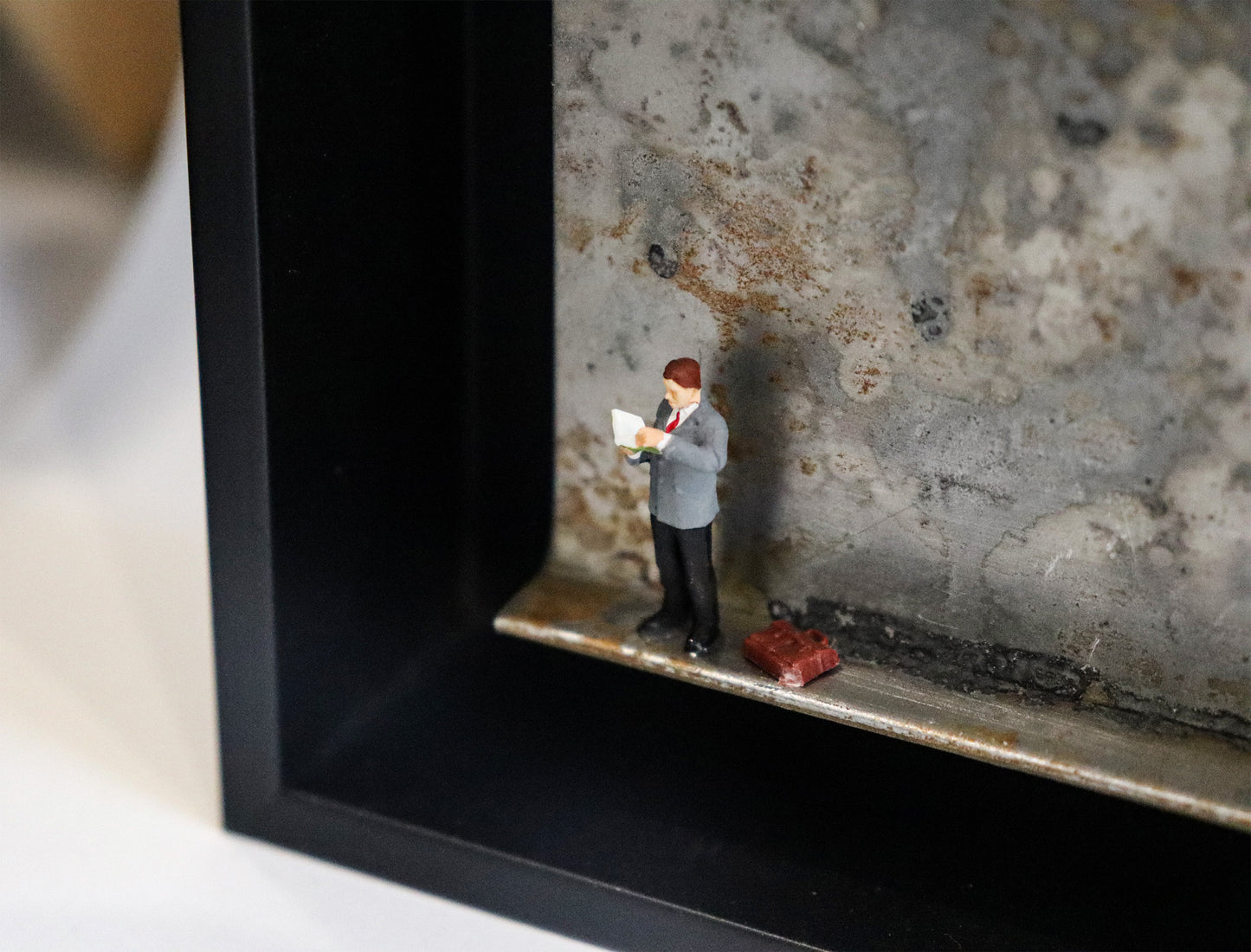 Original Objekt - Assemblage von Katrin Schramm, Titel „Bücher sind Lebensmittel“ – Metallwinkel mit Miniatur-Figur, Detailansicht: Ein Mann im Anzug steht am linken Rand des Winkels. Er schaut in ein geöffnetes Buch. Neben ihm steht eine Reisetasche. Dahinter das Metall des Winkels mit Struktur, als Hintergrund. In einem schwarzen Rahmen aus Holz. Objektcollage, erhältlich bei Galerie Waldbach - What The Artist Made
