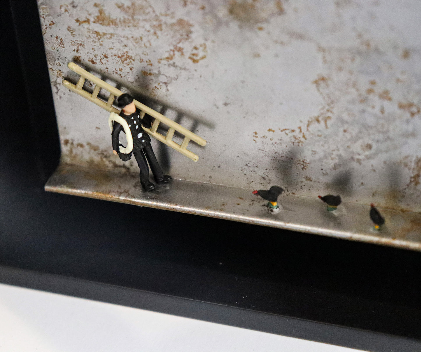 Original Objekt - Assemblage von Katrin Schramm, Titel „Das Glück ist schwarz“ – Metallwinkel mit Miniatur-Figuren, Detailansicht: Ein Schornsteinfeger steht mit Leiter und Seil steht am linken Rand des Winkels. Rechts von ihm drei schwarze Hühner. Dahinter das Metall des Winkels mit Struktur, als Hintergrund. In einem schwarzen Rahmen aus Holz. Objektcollage, erhältlich bei Galerie Waldbach - What The Artist Made