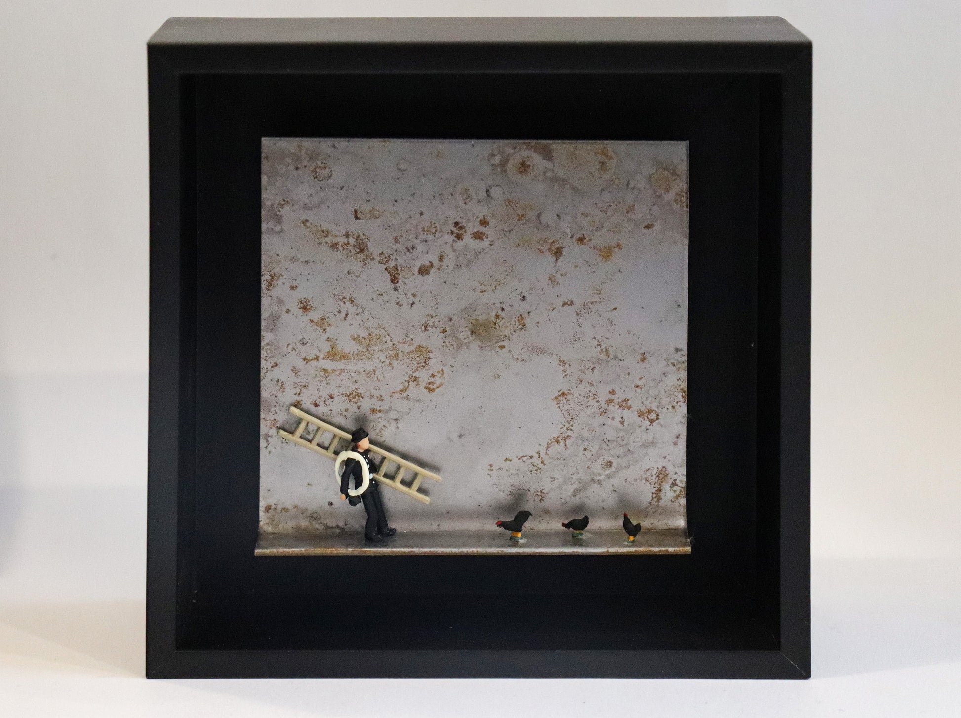 Original Objekt - Assemblage von Katrin Schramm, Titel „Das Glück ist schwarz“ – Metallwinkel mit Miniatur-Figuren: Ein Schornsteinfeger steht mit Leiter und Seil steht am linken Rand des Winkels. Rechts von ihm drei schwarze Hühner. Dahinter das Metall des Winkels mit Struktur, als Hintergrund. In einem schwarzen Rahmen aus Holz. Objektcollage, erhältlich bei Galerie Waldbach - What The Artist Made