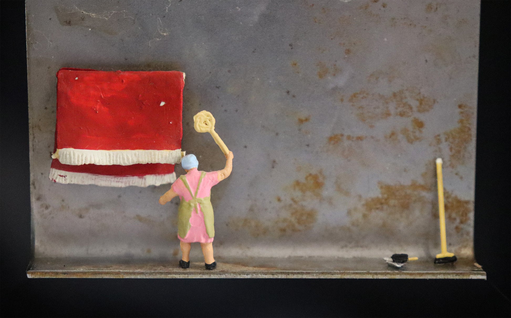 Original Objekt - Assemblage von Katrin Schramm, Titel „Das bisschen Haushalt“ – Metallwinkel mit Miniatur-Figur, Detailansicht: Eine Frau in Haushaltskleidung klopft einen am Metallwinkel hängenden roten Teppich aus. Am rechten Bildrand lehnt ein Kehrbesen am Metall und ein Kehrblech liegt daneben. Dahinter das Metall des Winkels mit Struktur, als Hintergrund. In einem schwarzen Rahmen aus Holz. Objektcollage, erhältlich bei Galerie Waldbach - What The Artist Made