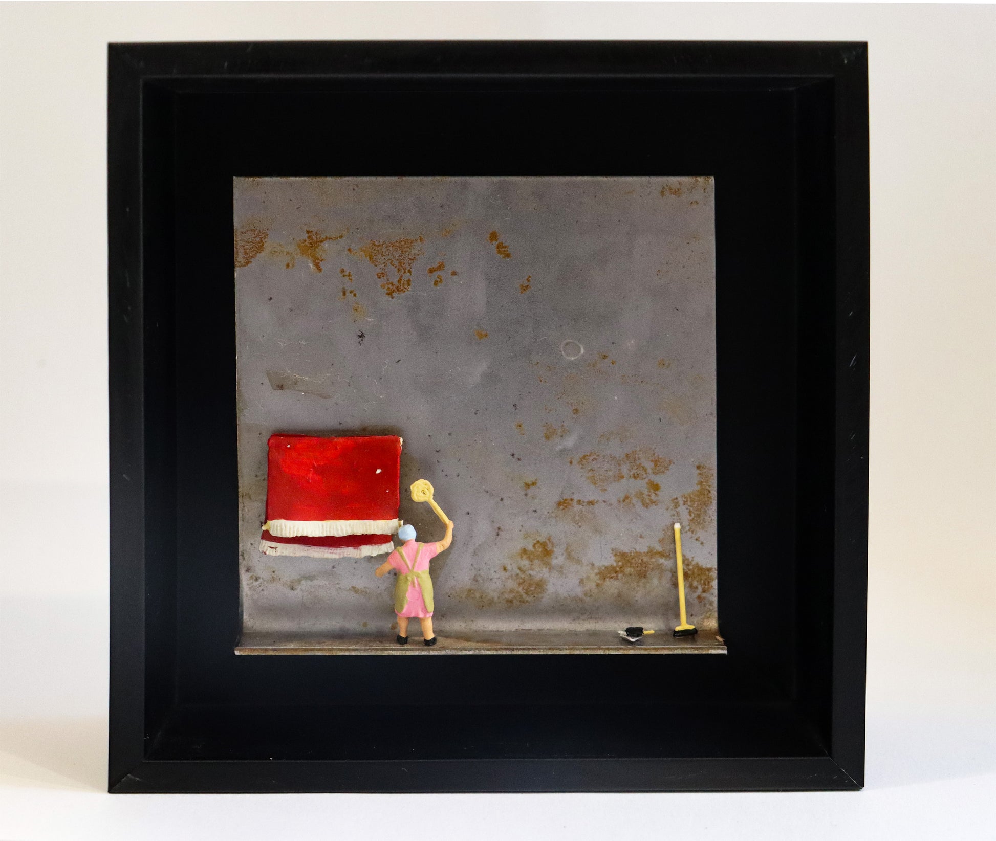 Original Objekt - Assemblage von Katrin Schramm, Titel „Das bisschen Haushalt“ – Metallwinkel mit Miniatur-Figur: Eine Frau in Haushaltskleidung klopft einen am Metallwinkel hängenden roten Teppich aus. Am rechten Bildrand lehnt ein Kehrbesen am Metall und ein Kehrblech liegt daneben. Dahinter das Metall des Winkels mit Struktur, als Hintergrund. In einem schwarzen Rahmen aus Holz. Objektcollage, erhältlich bei Galerie Waldbach - What The Artist Made