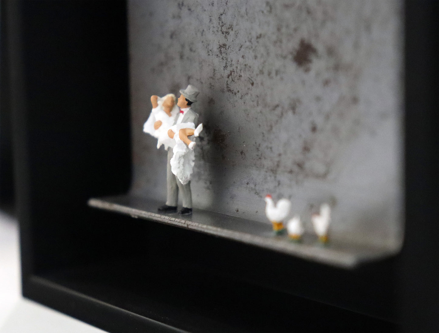 Original Objekt - Assemblage von Katrin Schramm, Titel „Dem Himmel so nah...“ – Metallwinkel mit Miniatur-Figuren. Detailansicht vom Brautpaar. Dahinter das Metall des Winkels mit Struktur. Objektcollage im schwarzen Rahmen, erhältlich bei Galerie Waldbach - What The Artist Made