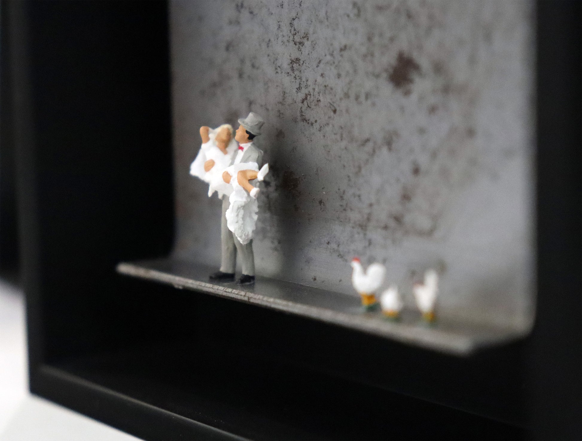 Original Objekt - Assemblage von Katrin Schramm, Titel „Dem Himmel so nah...“ – Metallwinkel mit Miniatur-Figuren. Detailansicht vom Brautpaar. Dahinter das Metall des Winkels mit Struktur. Objektcollage im schwarzen Rahmen, erhältlich bei Galerie Waldbach - What The Artist Made