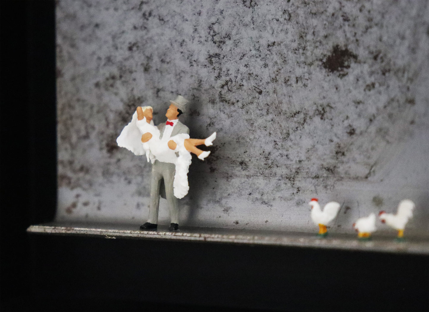 Original Objekt - Assemblage von Katrin Schramm, Titel „Dem Himmel so nah...“ – Metallwinkel mit Miniatur-Figuren. Detailansicht vom Brautpaar. Dahinter das Metall des Winkels mit Struktur. Objektcollage im schwarzen Rahmen, erhältlich bei Galerie Waldbach - What The Artist Made