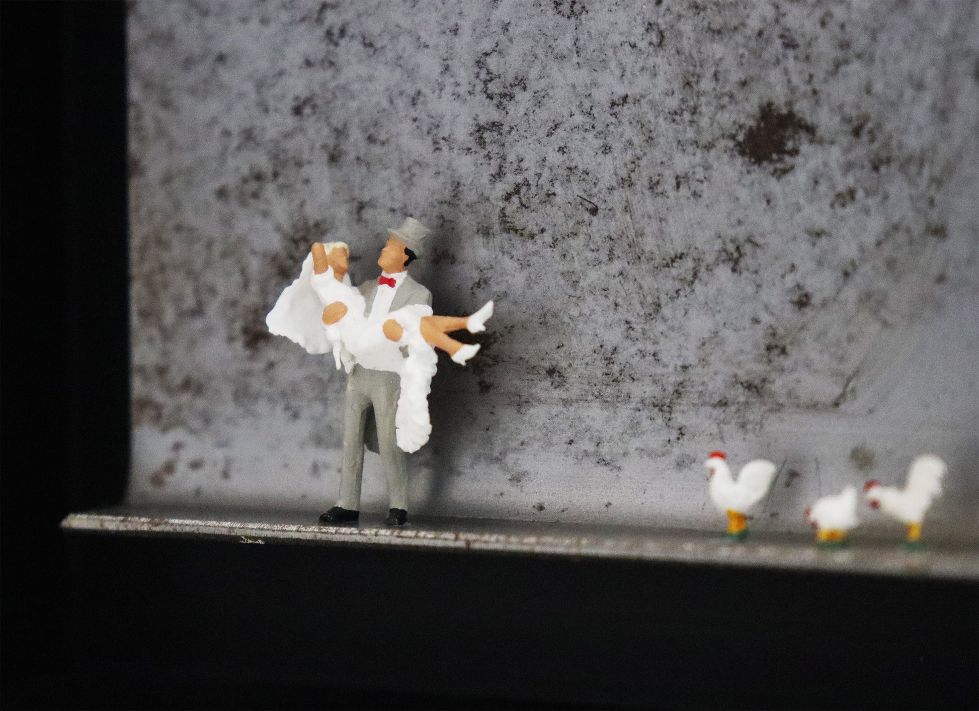 Original Objekt - Assemblage von Katrin Schramm, Titel „Dem Himmel so nah...“ – Metallwinkel mit Miniatur-Figuren. Detailansicht vom Brautpaar. Dahinter das Metall des Winkels mit Struktur. Objektcollage im schwarzen Rahmen, erhältlich bei Galerie Waldbach - What The Artist Made