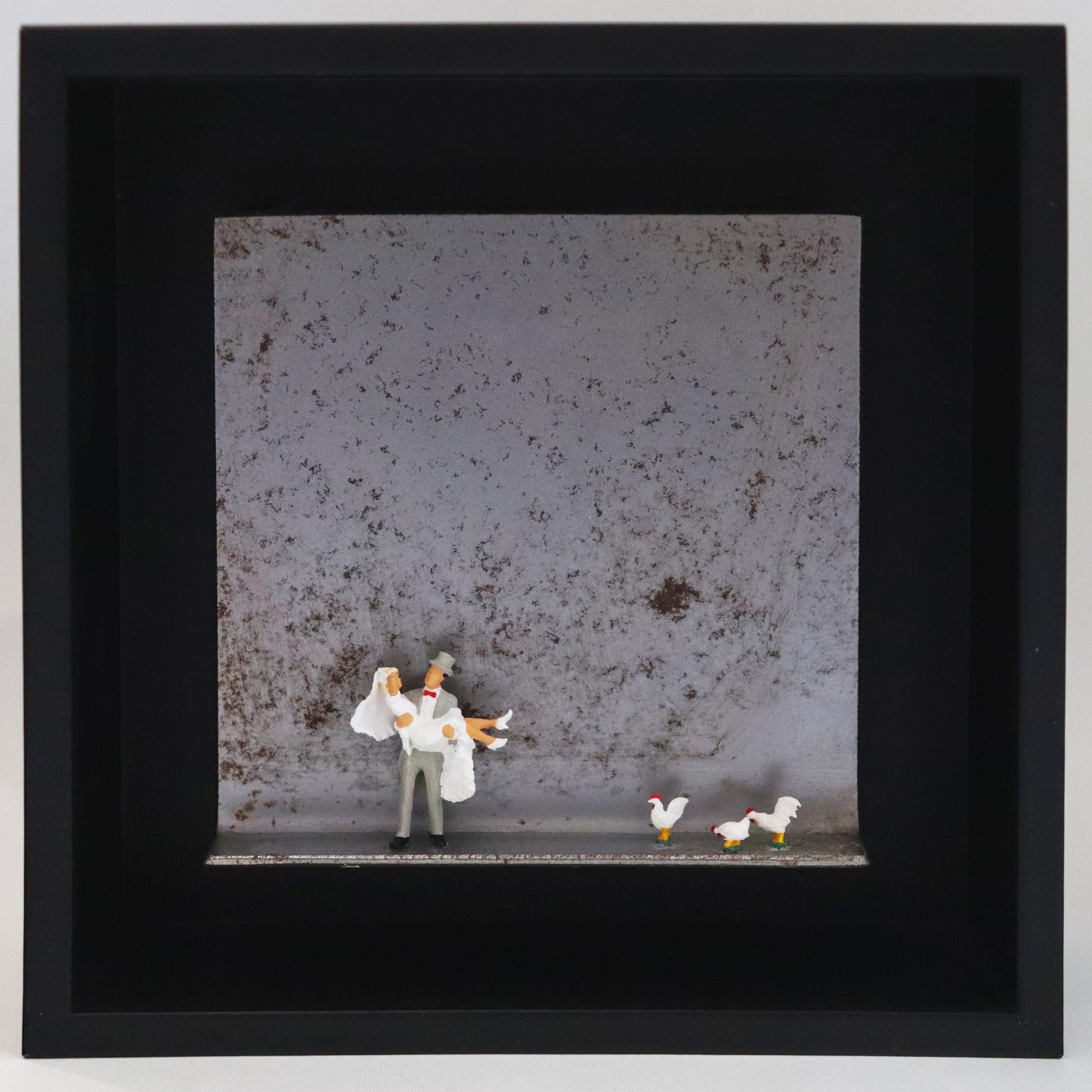 Original Objekt - Assemblage von Katrin Schramm, Titel „Dem Himmel so nah...“ – Metallwinkel mit Miniatur-Figuren: Ein Brautpaar, der Bräutigam trägt die Braut in den Armen. Rechts neben ihnen stehen drei weiße Hühner. Dahinter das Metall des Winkels mit Struktur, als Hintergrund. Objektcollage im schwarzen Rahmen, erhältlich bei Galerie Waldbach - What The Artist Made