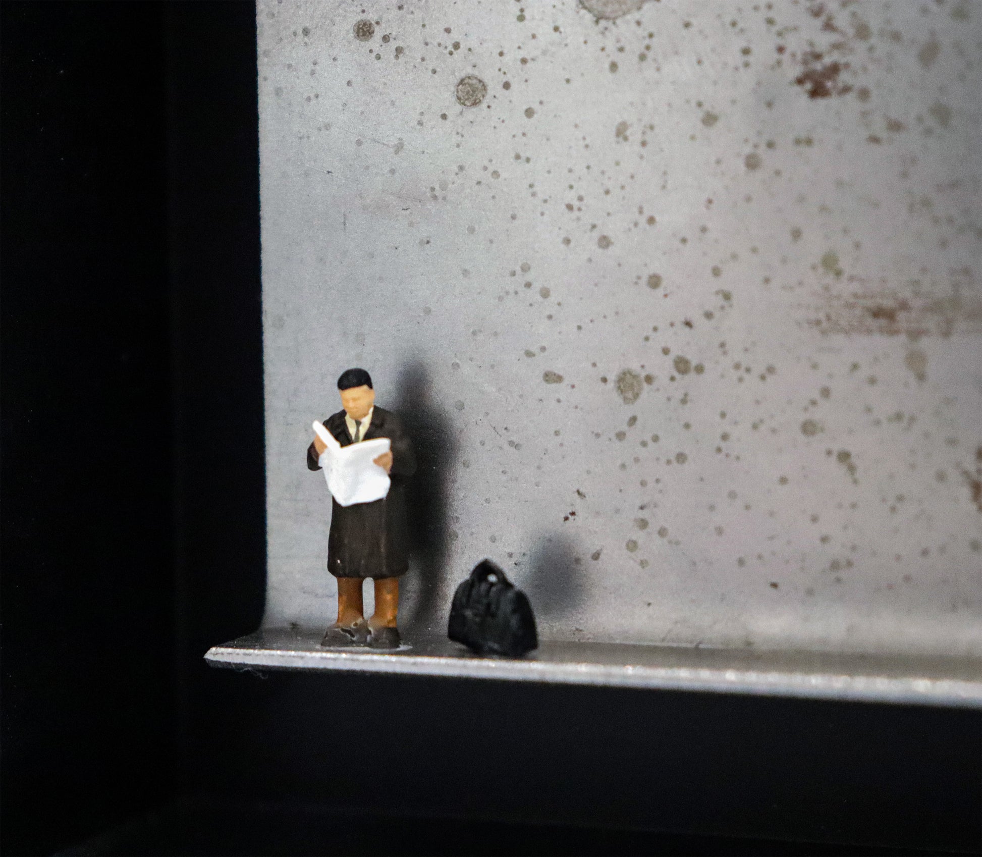 Original Objekt - Assemblage von Katrin Schramm, Titel „Der letzte Zeitungsleser?“ – Metallwinkel mit Miniatur-Figur, Detailansicht. Dahinter das Metall des Winkels mit Struktur, als Hintergrund. Objektcollage im schwarzen Rahmen, erhältlich bei Galerie Waldbach - What The Artist Made
