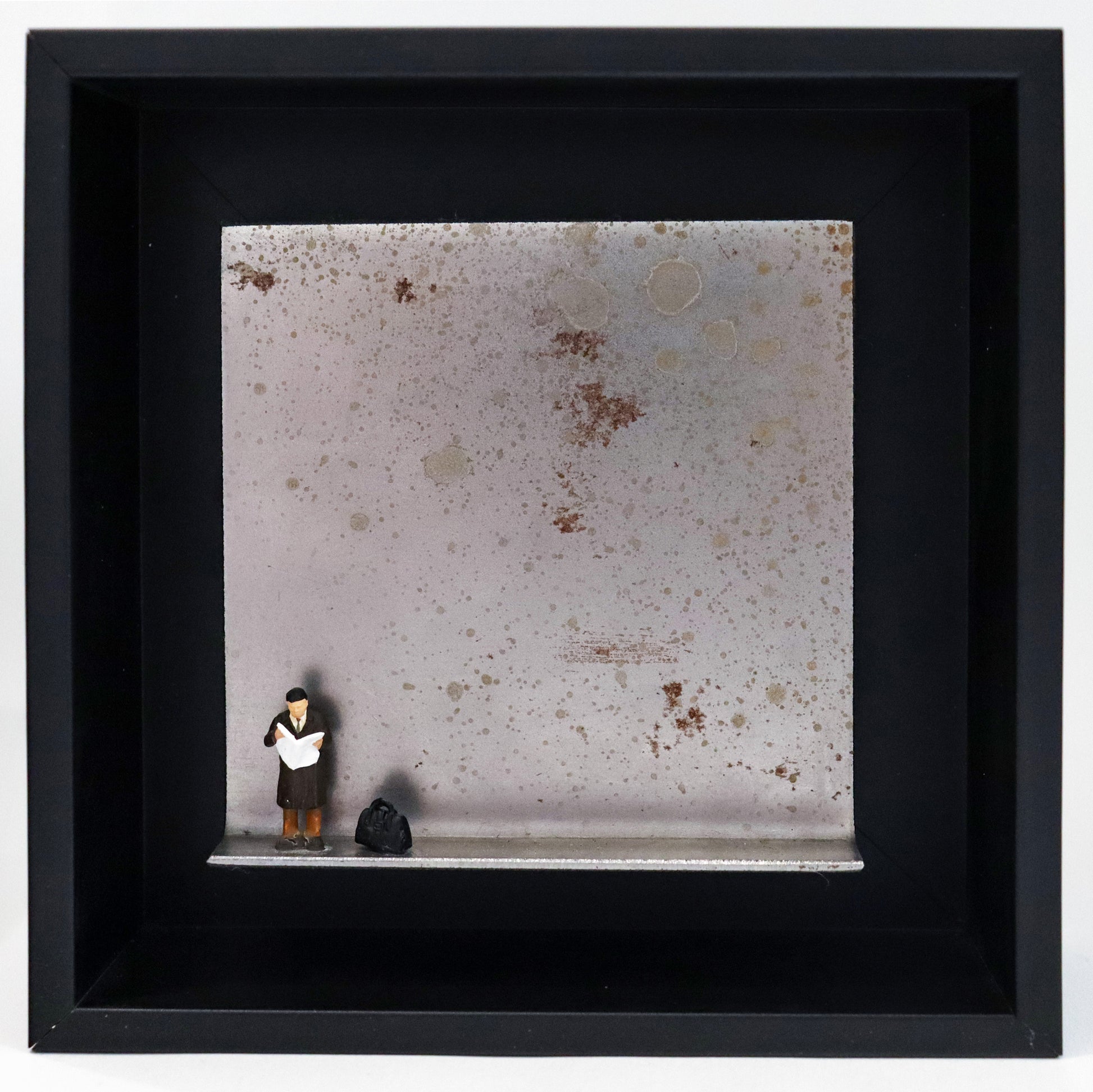 Original Objekt - Assemblage von Katrin Schramm, Titel „Der letzte Zeitungsleser?“ – Metallwinkel mit Miniatur-Figur: Ein Mann steht am linken Rand des Winkels und liest eine Zeitung. Neben ihm steht eine schwarze Reisetasche. Dahinter das Metall des Winkels mit Struktur, als Hintergrund. Objektcollage im schwarzen Rahmen, erhältlich bei Galerie Waldbach - What The Artist Made