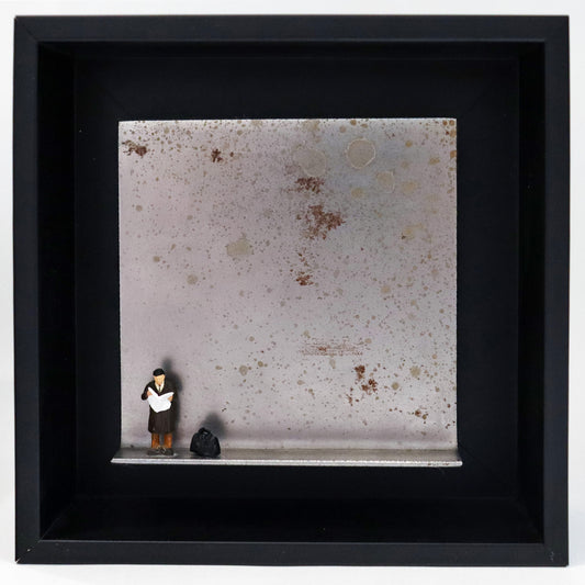 Original Objekt - Assemblage von Katrin Schramm, Titel „Der letzte Zeitungsleser?“ – Metallwinkel mit Miniatur-Figur: Ein Mann steht am linken Rand des Winkels und liest eine Zeitung. Neben ihm steht eine schwarze Reisetasche. Dahinter das Metall des Winkels mit Struktur, als Hintergrund. Objektcollage im schwarzen Rahmen, erhältlich bei Galerie Waldbach - What The Artist Made