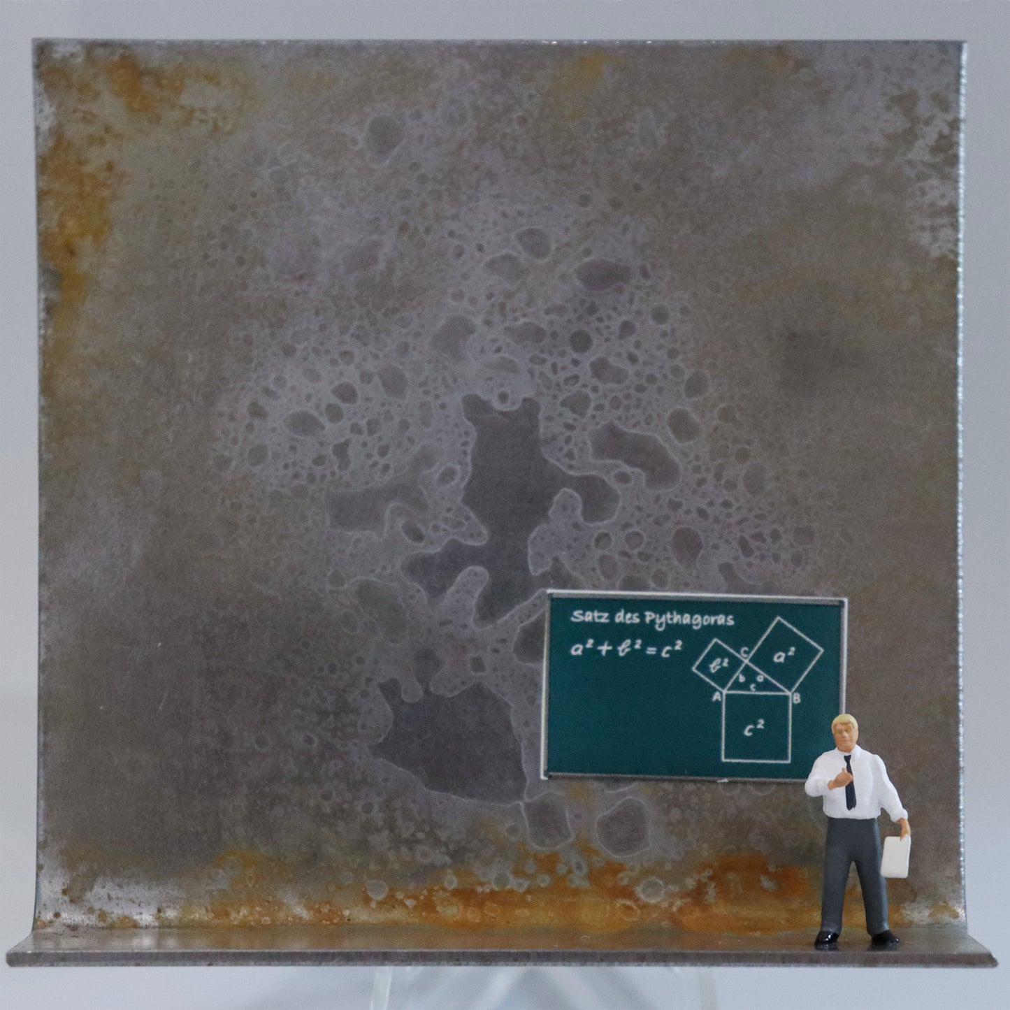 Original Objekt - Assemblage von Katrin Schramm, Titel „Die Zahl ist das Wesen aller Dinge.“  – Metallwinkel mit Miniatur-Figur: Ein Mann steht am rechten Rand des Winkels in Hemd und Krawatte und hält ein  Blatt in der Hand. Hinter ihm ist eine Schultafel, die den Satz des Pythagoras zeigt. Dahinter das Metall des Winkels mit Struktur, als Hintergrund. Objektcollage, erhältlich bei Galerie Waldbach - What The Artist Made