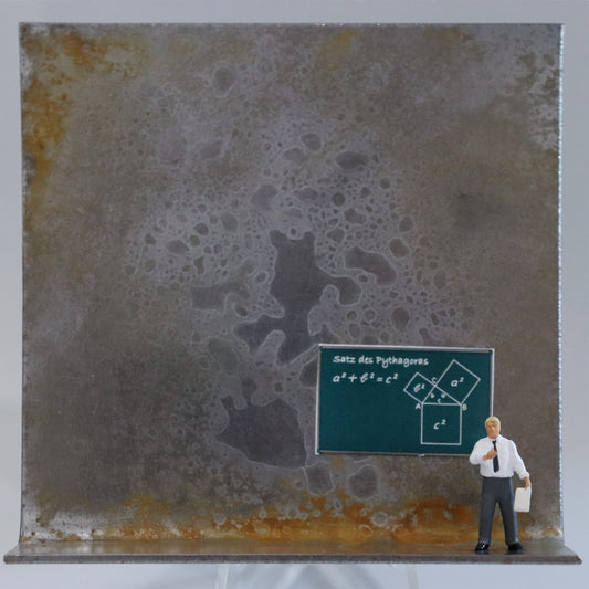 Original Objekt - Assemblage von Katrin Schramm, Titel „Die Zahl ist das Wesen aller Dinge.“  – Metallwinkel mit Miniatur-Figur: Ein Mann steht am rechten Rand des Winkels in Hemd und Krawatte und hält ein  Blatt in der Hand. Hinter ihm ist eine Schultafel, die den Satz des Pythagoras zeigt. Dahinter das Metall des Winkels mit Struktur, als Hintergrund. Objektcollage, erhältlich bei Galerie Waldbach - What The Artist Made