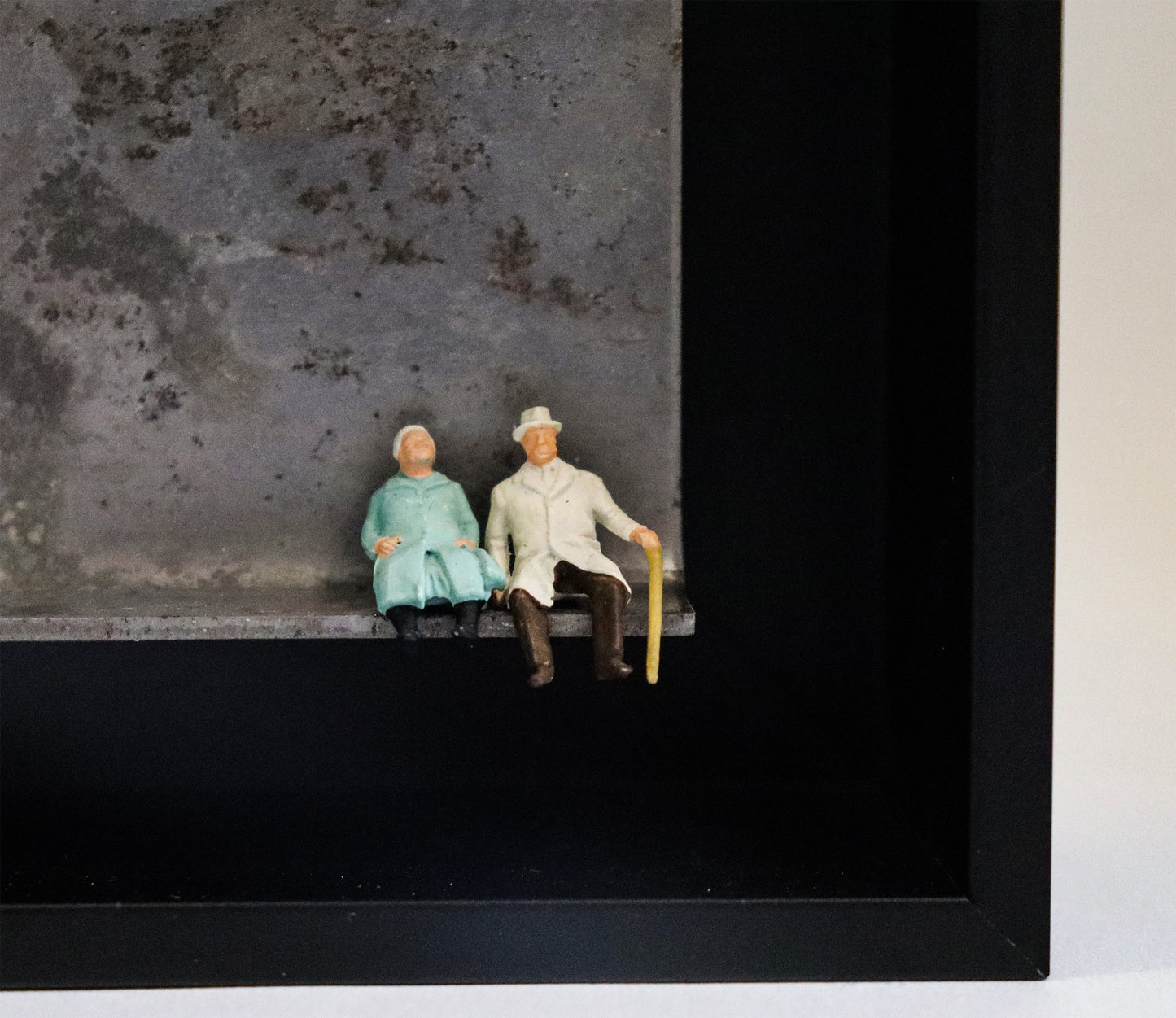 Original Objekt - Assemblage von Katrin Schramm, Titel „Durch dick und dünn“ – Metallwinkel mit Miniatur-Figuren, Detailansicht: Ein altes Paar sitzt am rechten Rand des Winkels, der Mann trägt einen Gehstock. Dahinter das Metall des Winkels mit Struktur, als Hintergrund. In einem schwarzen Rahmen aus Holz. Objektcollage, erhältlich bei Galerie Waldbach - What The Artist Made