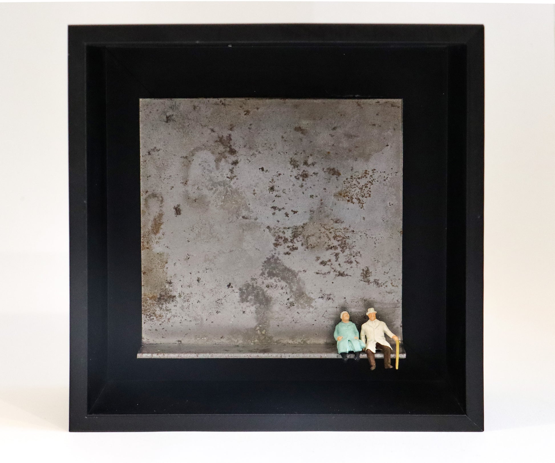 Original Objekt - Assemblage von Katrin Schramm, Titel „Durch dick und dünn“ – Metallwinkel mit Miniatur-Figuren: Ein altes Paar sitzt am rechten Rand des Winkels, der Mann trägt einen Gehstock. Dahinter das Metall des Winkels mit Struktur, als Hintergrund. In einem schwarzen Rahmen aus Holz. Objektcollage, erhältlich bei Galerie Waldbach - What The Artist Made