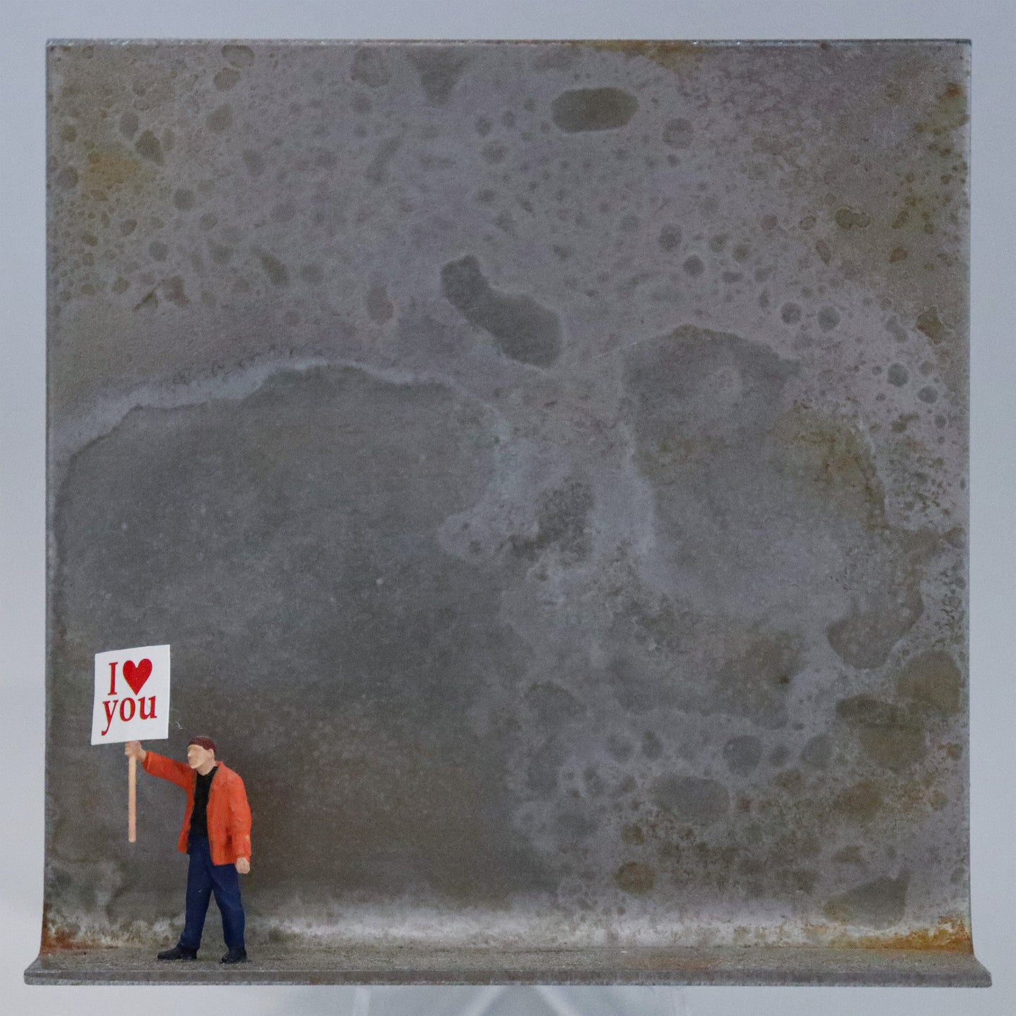 Original Objekt - Assemblage von Katrin Schramm, Titel „Einfach so!“  – Metallwinkel mit Miniatur-Figur: Eine Person in orangener Jacke steht am linken Rand des Winkels und hält ein Schild hoch. Darauf steht "I (heart) you". Dahinter das Metall des Winkels mit Struktur, als Hintergrund. Objektcollage, erhältlich bei Galerie Waldbach - What The Artist Made