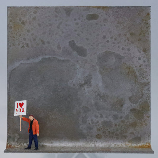 Original Objekt - Assemblage von Katrin Schramm, Titel „Einfach so!“  – Metallwinkel mit Miniatur-Figur: Eine Person in orangener Jacke steht am linken Rand des Winkels und hält ein Schild hoch. Darauf steht "I (heart) you". Dahinter das Metall des Winkels mit Struktur, als Hintergrund. Objektcollage, erhältlich bei Galerie Waldbach - What The Artist Made