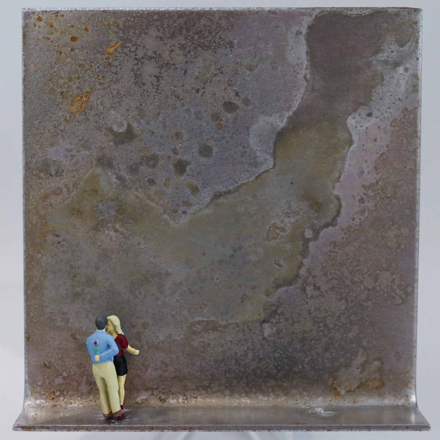Original Objekt - Assemblage von Katrin Schramm, Titel „Endlich!“  – Metallwinkel mit Miniatur-Figuren: Ein Paar steht am linken Rand des Winkels. Der Mann mit einem Fuß auf einem Fuß der Frau stehend, hält er eine Rose hinter dem Rücken, während sie nah beieinander sind. Dahinter das Metall des Winkels mit Struktur, als Hintergrund. Objektcollage, erhältlich bei Galerie Waldbach - What The Artist Made