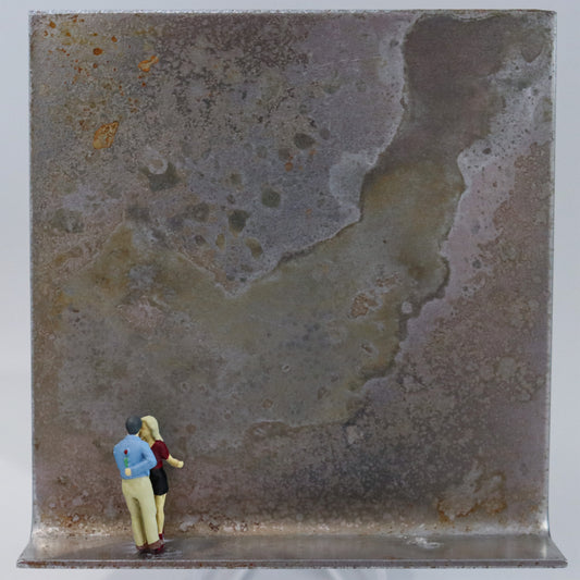 Original Objekt - Assemblage von Katrin Schramm, Titel „Endlich!“  – Metallwinkel mit Miniatur-Figuren: Ein Paar steht am linken Rand des Winkels. Der Mann mit einem Fuß auf einem Fuß der Frau stehend, hält er eine Rose hinter dem Rücken, während sie nah beieinander sind. Dahinter das Metall des Winkels mit Struktur, als Hintergrund. Objektcollage, erhältlich bei Galerie Waldbach - What The Artist Made