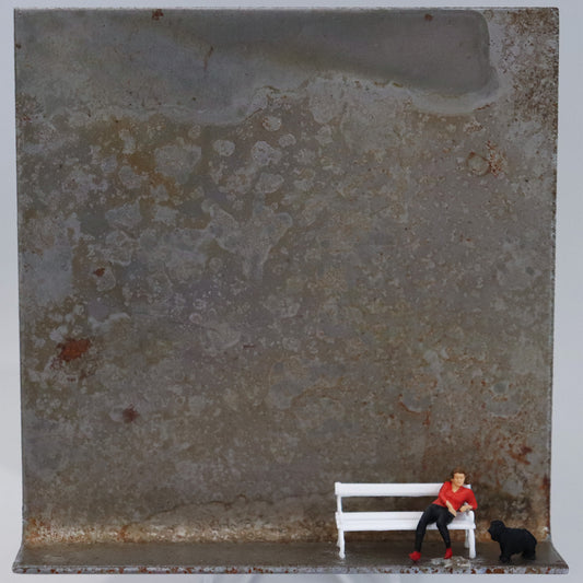 Original Objekt - Assemblage von Katrin Schramm, Titel „Gelassen.“  – Metallwinkel mit Miniatur-Figuren: Eine Frau in rot-schwarzer Kleidung sitzt am rechten Rand des Winkels auf einer weißen Bank. Neben ihr steht ein schwarzer Hund. Dahinter das Metall des Winkels mit Struktur, als Hintergrund. Objektcollage, erhältlich bei Galerie Waldbach - What The Artist Made