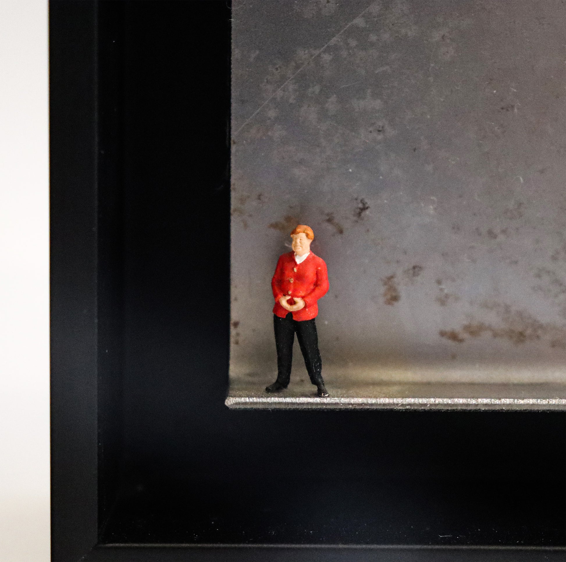 Original Objekt - Assemblage von Katrin Schramm, Titel „Geschafft“ – Metallwinkel mit Miniatur-Figur, Detailansicht: Eine Person, dem Erscheinen nach soll sie Angela Merkel darstellen, steht am linken Rand des Winkels. Sie trägt einen roten Blazer und  hat die Hände zur typischen Raute geformt. Dahinter das Metall des Winkels mit Struktur, als Hintergrund. In einem schwarzen Rahmen aus Holz. Objektcollage, erhältlich bei Galerie Waldbach - What The Artist Made