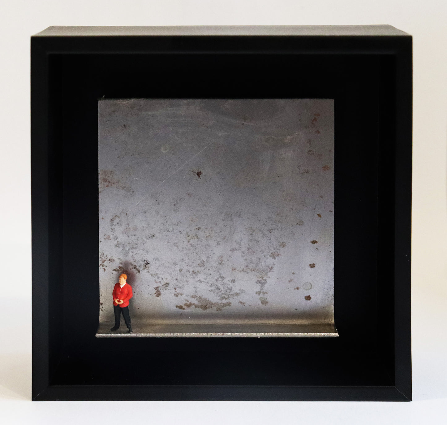 Original Objekt - Assemblage von Katrin Schramm, Titel „Geschafft“ – Metallwinkel mit Miniatur-Figur: Eine Person, dem Erscheinen nach soll sie Angela Merkel darstellen, steht am linken Rand des Winkels. Sie trägt einen roten Blazer und  hat die Hände zur typischen Raute geformt. Dahinter das Metall des Winkels mit Struktur, als Hintergrund. In einem schwarzen Rahmen aus Holz. Objektcollage, erhältlich bei Galerie Waldbach - What The Artist Made