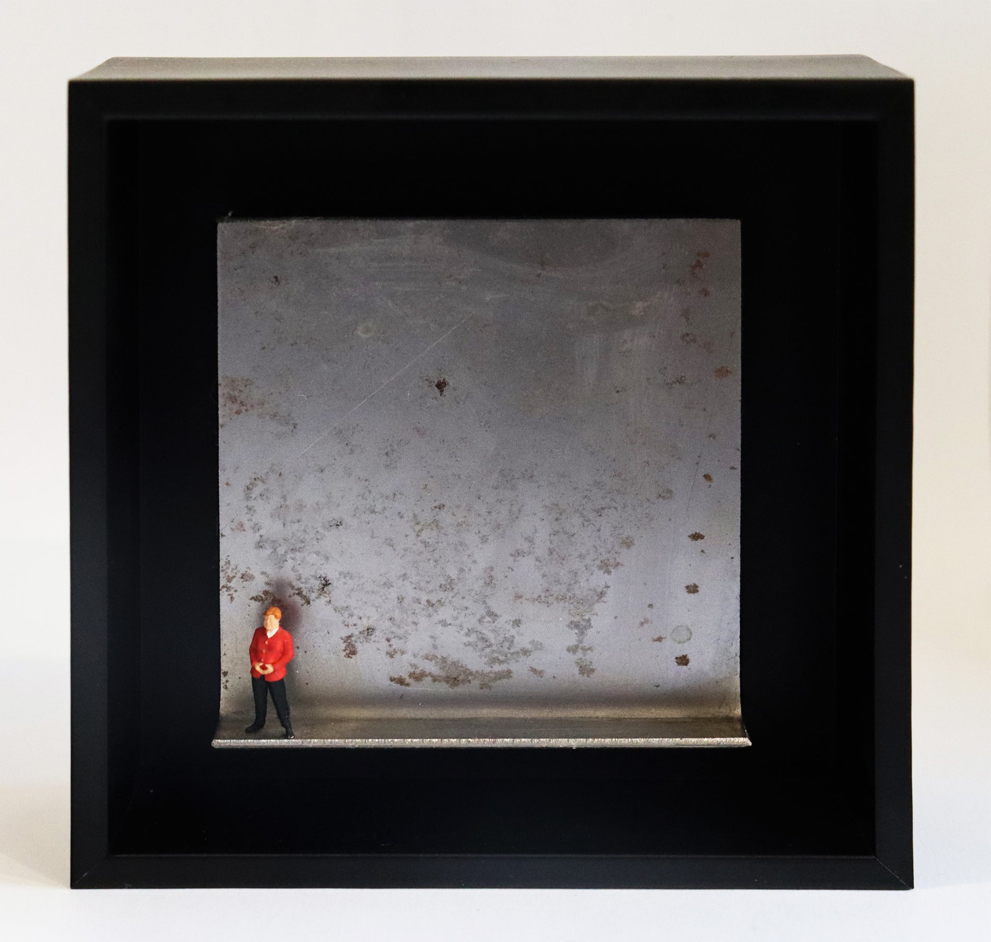 Original Objekt - Assemblage von Katrin Schramm, Titel „Geschafft“ – Metallwinkel mit Miniatur-Figur: Eine Person, dem Erscheinen nach soll sie Angela Merkel darstellen, steht am linken Rand des Winkels. Sie trägt einen roten Blazer und  hat die Hände zur typischen Raute geformt. Dahinter das Metall des Winkels mit Struktur, als Hintergrund. In einem schwarzen Rahmen aus Holz. Objektcollage, erhältlich bei Galerie Waldbach - What The Artist Made