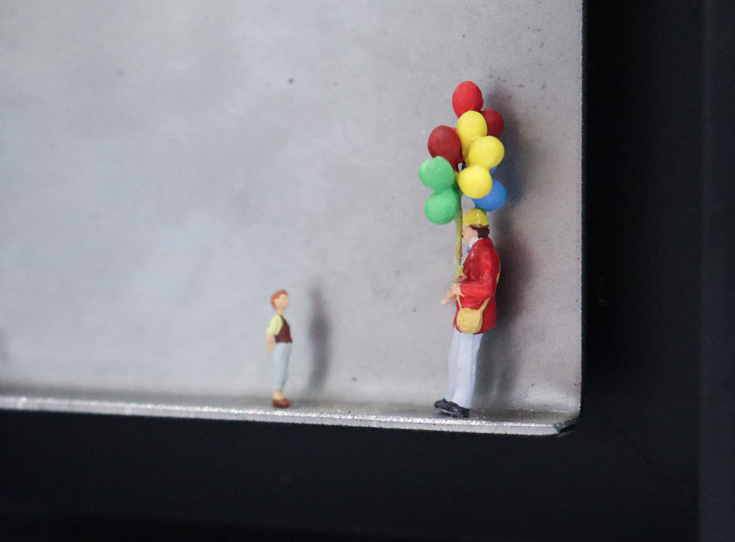 Original Objekt - Assemblage von Katrin Schramm, Titel „Lass sie fliegen...“ – Metallwinkel mit Miniatur-Figuren: Ein Ballonverkäufer steht auf der rechten Seite des Winkels und hält ein Bündel Ballons in der Hand. Vor ihm steht ein Kind und schaut  zu ihm und den Ballons hoch. Dahinter das Metall des Winkels mit Struktur, als Hintergrund. Objektcollage im schwarzen Rahmen, erhältlich bei Galerie Waldbach - What The Artist Made