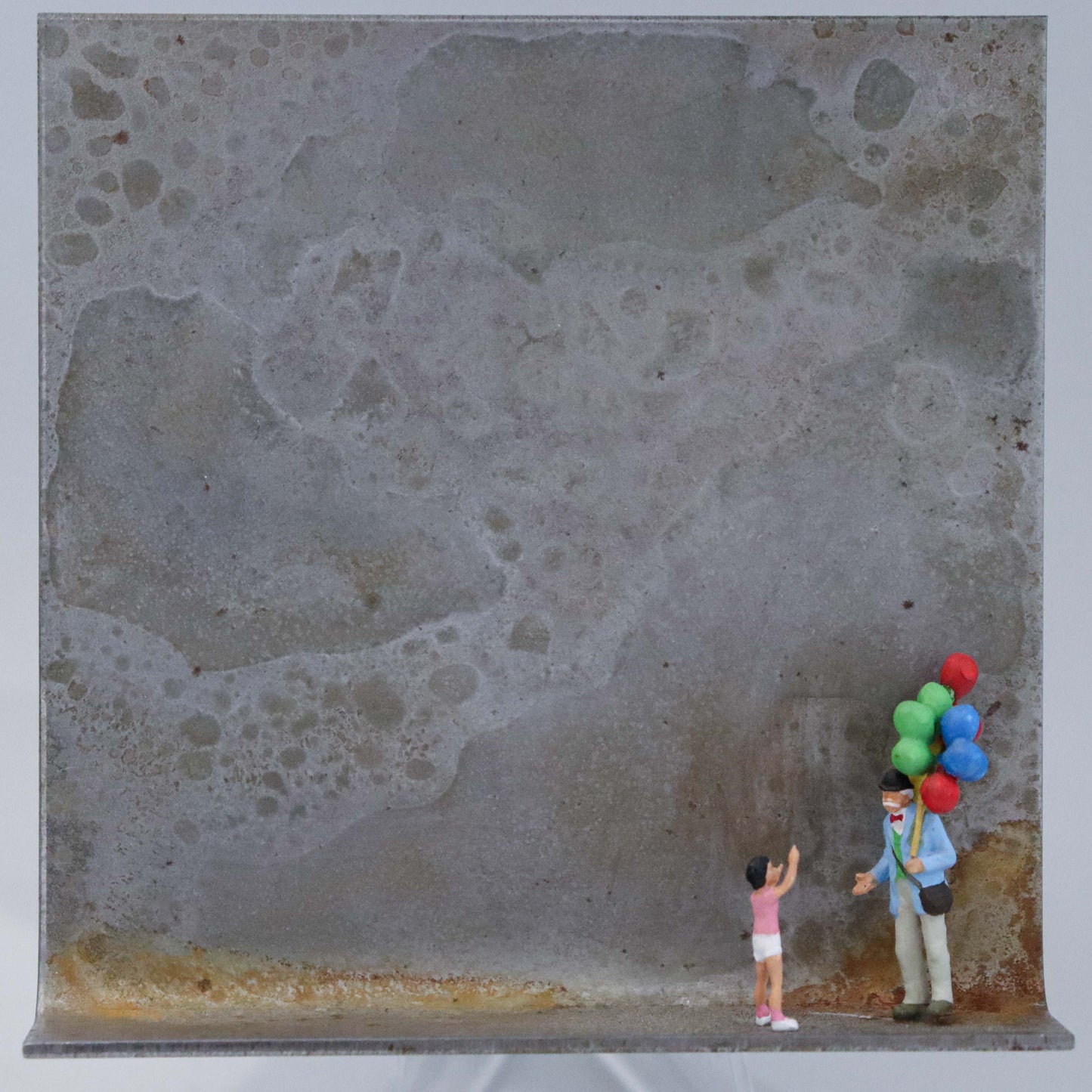 Original Objekt - Assemblage von Katrin Schramm, Titel „Lass sie fliegen!“  – Metallwinkel mit Miniatur-Figuren: Ein Kind zeigt auf die Luftballons eines Luftballonverkäufers mit vielen Ballons. Beide stehen am rechten Rand des Winkels. Dahinter das Metall des Winkels mit Struktur, als Hintergrund. Objektcollage, erhältlich bei Galerie Waldbach - What The Artist Made
