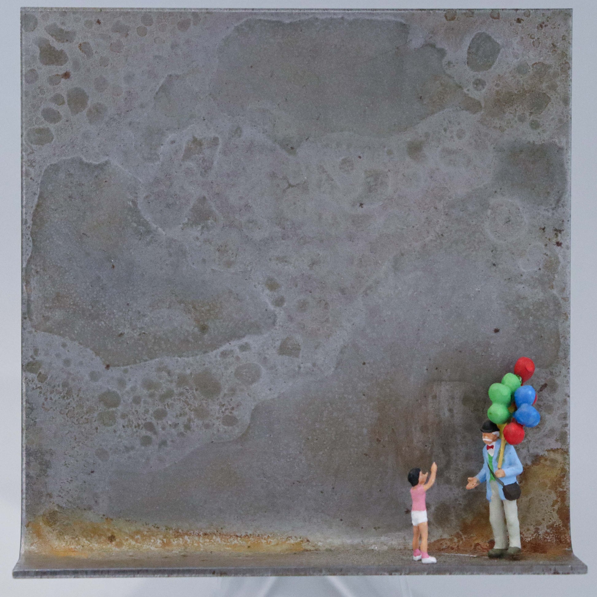 Original Objekt - Assemblage von Katrin Schramm, Titel „Lass sie fliegen!“  – Metallwinkel mit Miniatur-Figuren: Ein Kind zeigt auf die Luftballons eines Luftballonverkäufers mit vielen Ballons. Beide stehen am rechten Rand des Winkels. Dahinter das Metall des Winkels mit Struktur, als Hintergrund. Objektcollage, erhältlich bei Galerie Waldbach - What The Artist Made