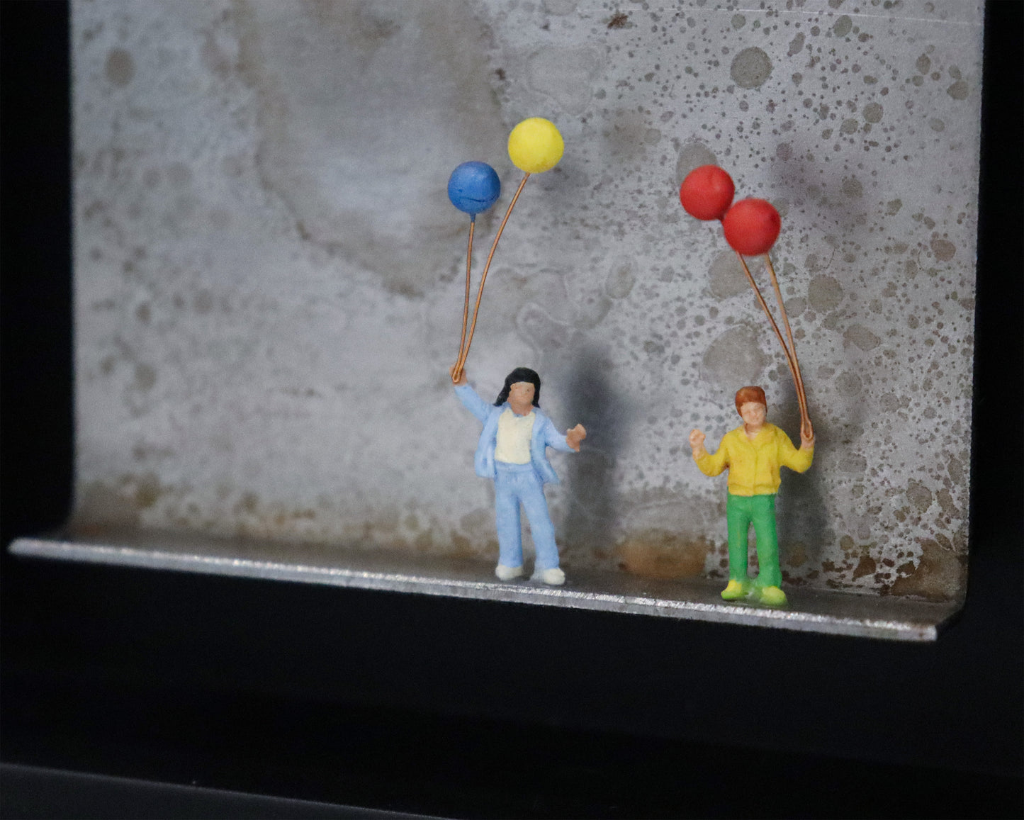 Original Objekt - Assemblage von Katrin Schramm, Titel „Mehr als nur Luft...“ – Metallwinkel mit Miniatur-Figuren, Detailansicht. Dahinter das Metall des Winkels mit Struktur, als Hintergrund. Objektcollage im schwarzen Rahmen, erhältlich bei Galerie Waldbach - What The Artist Made