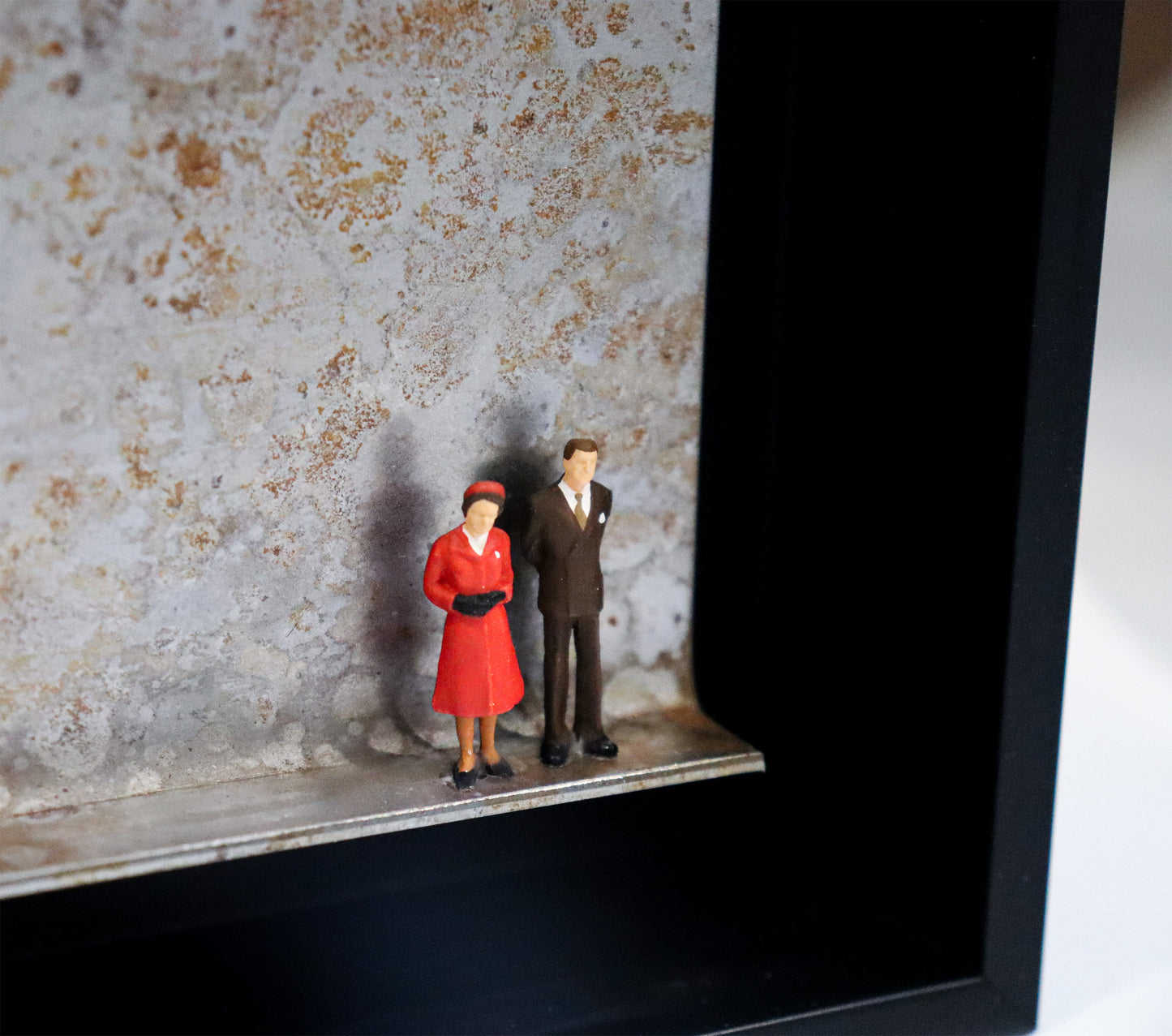 Original Objekt - Assemblage von Katrin Schramm, Titel „Never dressed down“ – Metallwinkel mit Miniatur-Figuren, Detailansicht:  Zwei Personen, Mann und Frau stehen an der rechten Seite des Winkels in formeller Kleidung, sie im roten Mantel, er im Anzug. Sie könnten die Queen und ihr Mann als jüngeres Paar sein. Dahinter das Metall des Winkels mit Struktur, als Hintergrund. In einem schwarzen Rahmen aus Holz. Objektcollage, erhältlich bei Galerie Waldbach - What The Artist Made