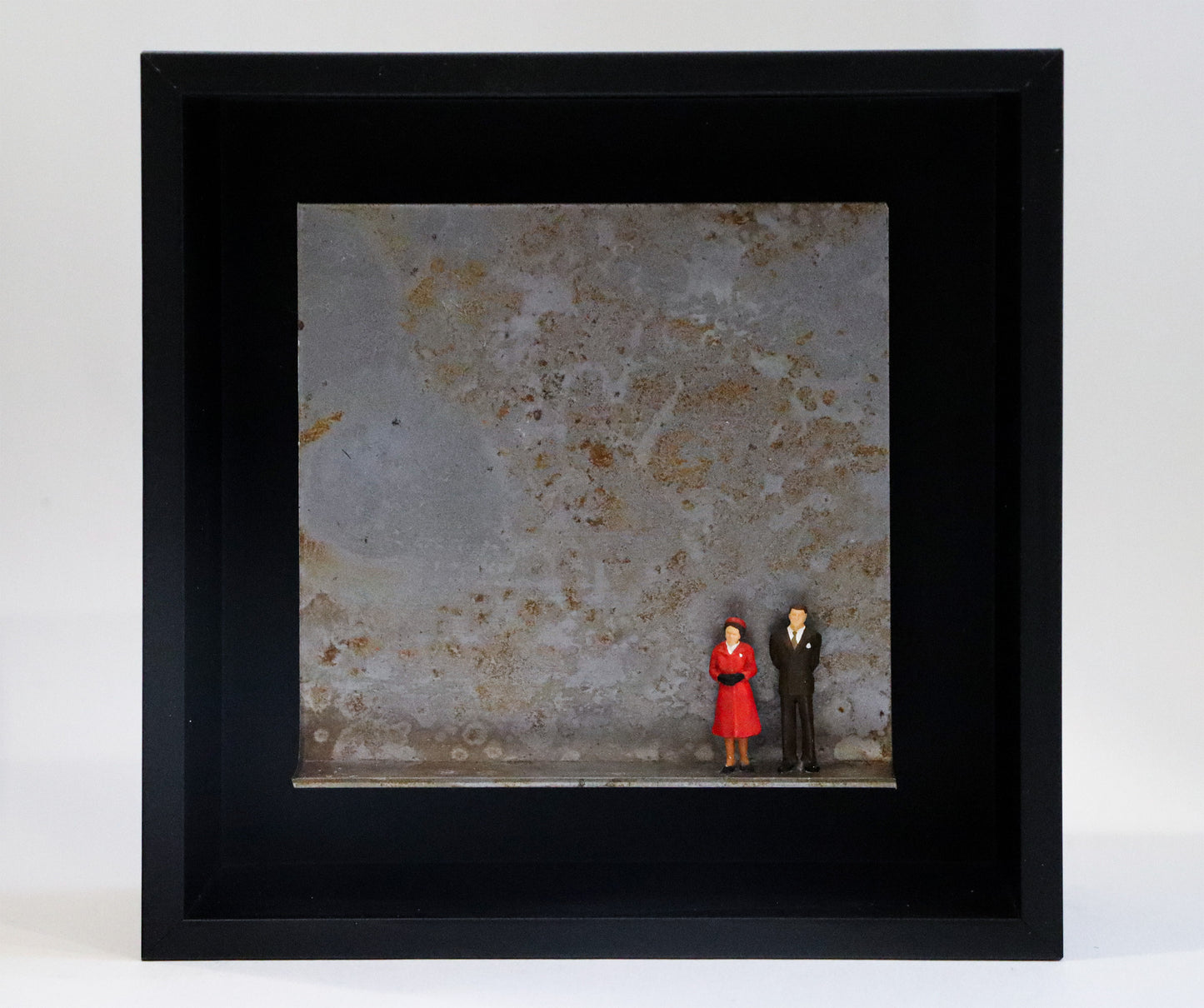 Original Objekt - Assemblage von Katrin Schramm, Titel „Never dressed down“ – Metallwinkel mit Miniatur-Figuren:  Zwei Personen, Mann und Frau stehen an der rechten Seite des Winkels in formeller Kleidung, sie im roten Mantel, er im Anzug. Sie könnten die Queen und ihr Mann als jüngeres Paar sein. Dahinter das Metall des Winkels mit Struktur, als Hintergrund. In einem schwarzen Rahmen aus Holz. Objektcollage, erhältlich bei Galerie Waldbach - What The Artist Made