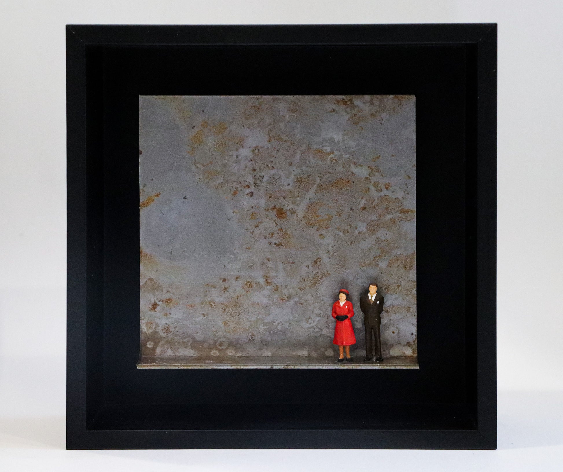 Original Objekt - Assemblage von Katrin Schramm, Titel „Never dressed down“ – Metallwinkel mit Miniatur-Figuren:  Zwei Personen, Mann und Frau stehen an der rechten Seite des Winkels in formeller Kleidung, sie im roten Mantel, er im Anzug. Sie könnten die Queen und ihr Mann als jüngeres Paar sein. Dahinter das Metall des Winkels mit Struktur, als Hintergrund. In einem schwarzen Rahmen aus Holz. Objektcollage, erhältlich bei Galerie Waldbach - What The Artist Made