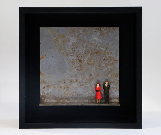 Original Objekt - Assemblage von Katrin Schramm, Titel „Never dressed down“ – Metallwinkel mit Miniatur-Figuren:  Zwei Personen, Mann und Frau stehen an der rechten Seite des Winkels in formeller Kleidung, sie im roten Mantel, er im Anzug. Sie könnten die Queen und ihr Mann als jüngeres Paar sein. Dahinter das Metall des Winkels mit Struktur, als Hintergrund. In einem schwarzen Rahmen aus Holz. Objektcollage, erhältlich bei Galerie Waldbach - What The Artist Made