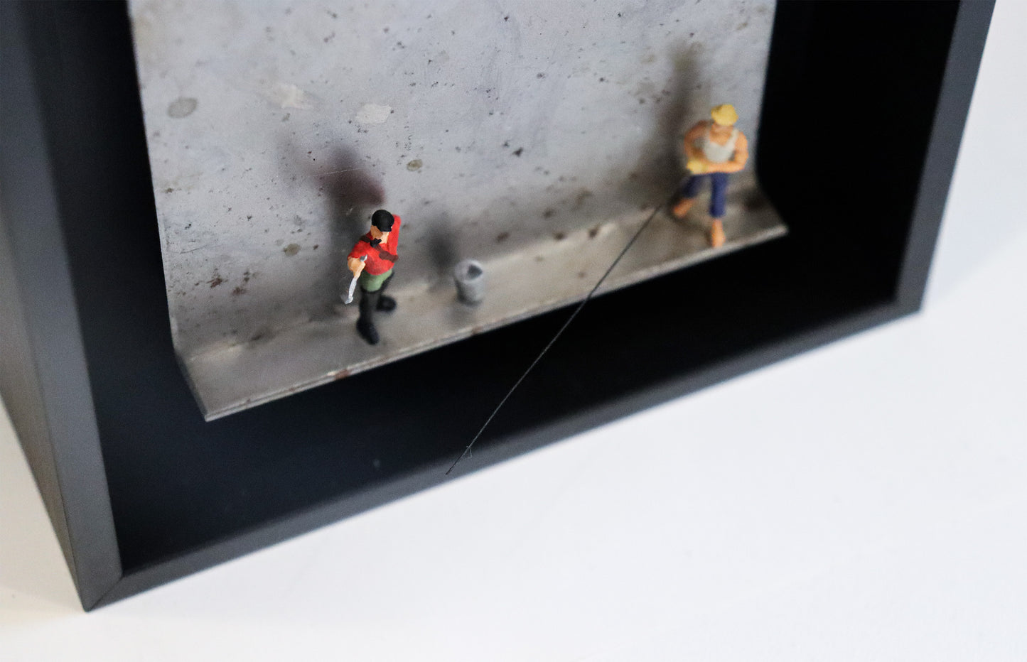 Original Objekt - Assemblage von Katrin Schramm, Titel „Petri Heil“ – Metallwinkel mit Miniatur-Figuren, Detail:  Zwei Angler stehen auf dem Winkel, einer hält einen Fisch hoch, neben ihm ein Eimer. Der andere hält eine Angel, die aus dem Winkel herausragt. Dahinter das Metall des Winkels mit Struktur, als Hintergrund. In einem schwarzen Rahmen aus Holz. Objektcollage, erhältlich bei Galerie Waldbach - What The Artist Made