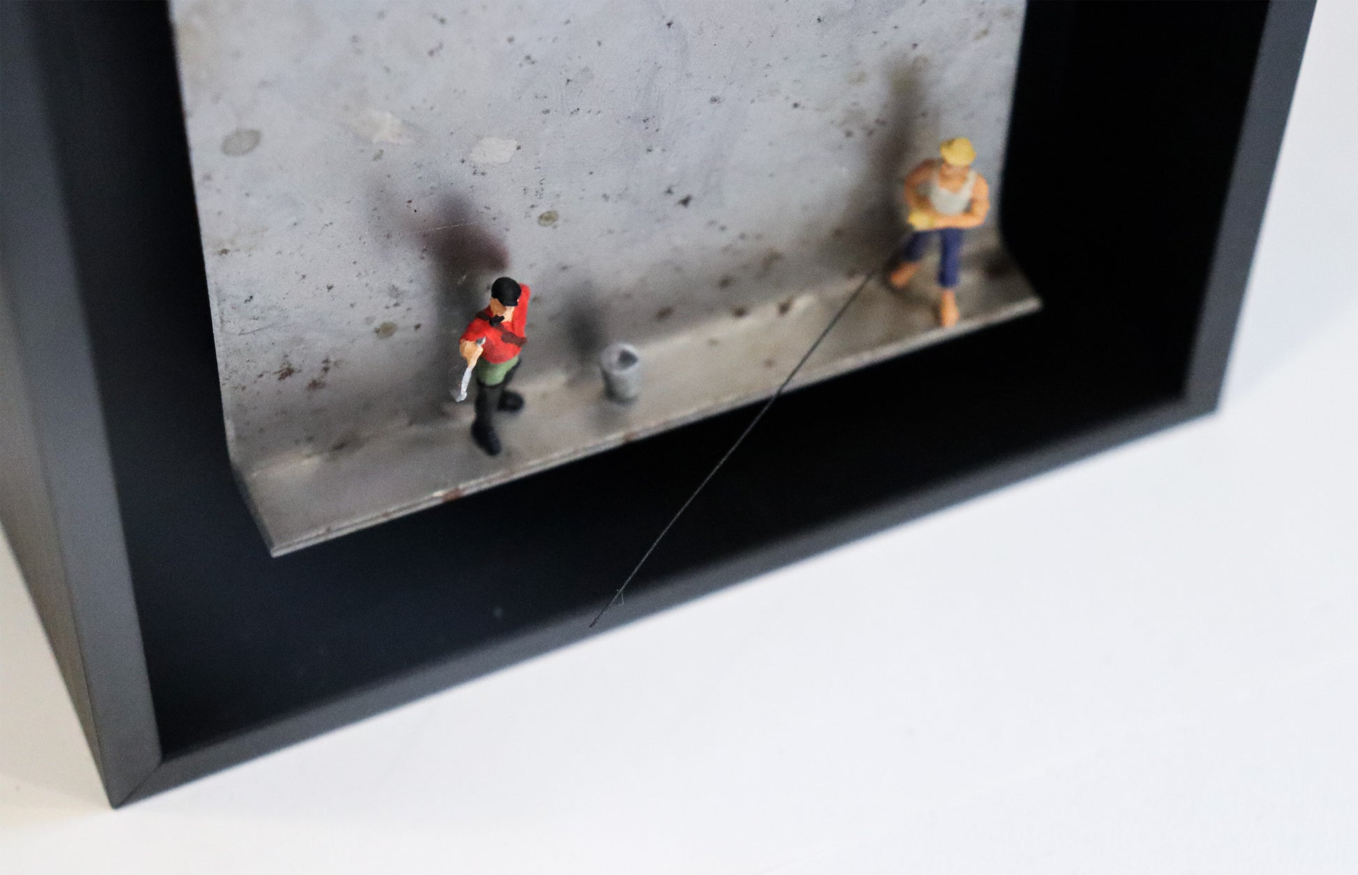 Original Objekt - Assemblage von Katrin Schramm, Titel „Petri Heil“ – Metallwinkel mit Miniatur-Figuren, Detail:  Zwei Angler stehen auf dem Winkel, einer hält einen Fisch hoch, neben ihm ein Eimer. Der andere hält eine Angel, die aus dem Winkel herausragt. Dahinter das Metall des Winkels mit Struktur, als Hintergrund. In einem schwarzen Rahmen aus Holz. Objektcollage, erhältlich bei Galerie Waldbach - What The Artist Made