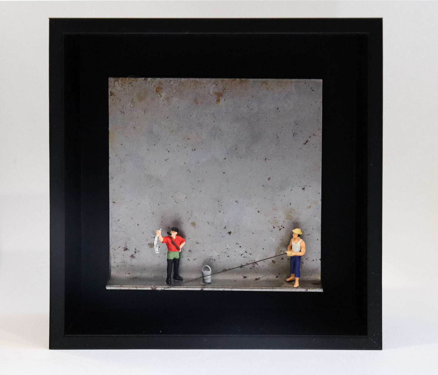 Original Objekt - Assemblage von Katrin Schramm, Titel „Petri Heil“ – Metallwinkel mit Miniatur-Figuren:  Zwei Angler stehen auf dem Winkel, einer hält einen Fisch hoch, neben ihm ein Eimer. Der andere hält eine Angel, die aus dem Winkel herausragt. Dahinter das Metall des Winkels mit Struktur, als Hintergrund. In einem schwarzen Rahmen aus Holz. Objektcollage, erhältlich bei Galerie Waldbach - What The Artist Made
