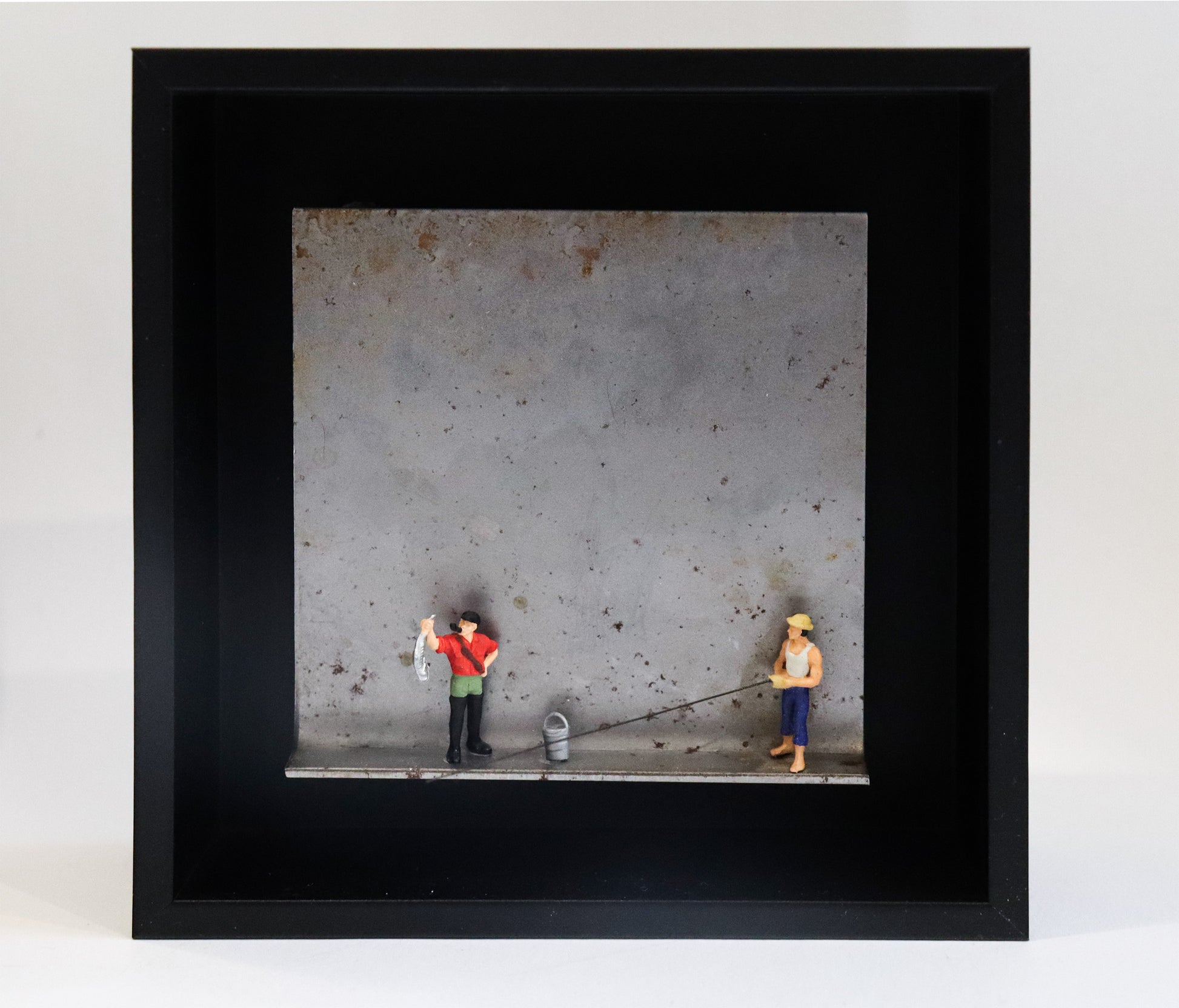 Original Objekt - Assemblage von Katrin Schramm, Titel „Petri Heil“ – Metallwinkel mit Miniatur-Figuren:  Zwei Angler stehen auf dem Winkel, einer hält einen Fisch hoch, neben ihm ein Eimer. Der andere hält eine Angel, die aus dem Winkel herausragt. Dahinter das Metall des Winkels mit Struktur, als Hintergrund. In einem schwarzen Rahmen aus Holz. Objektcollage, erhältlich bei Galerie Waldbach - What The Artist Made